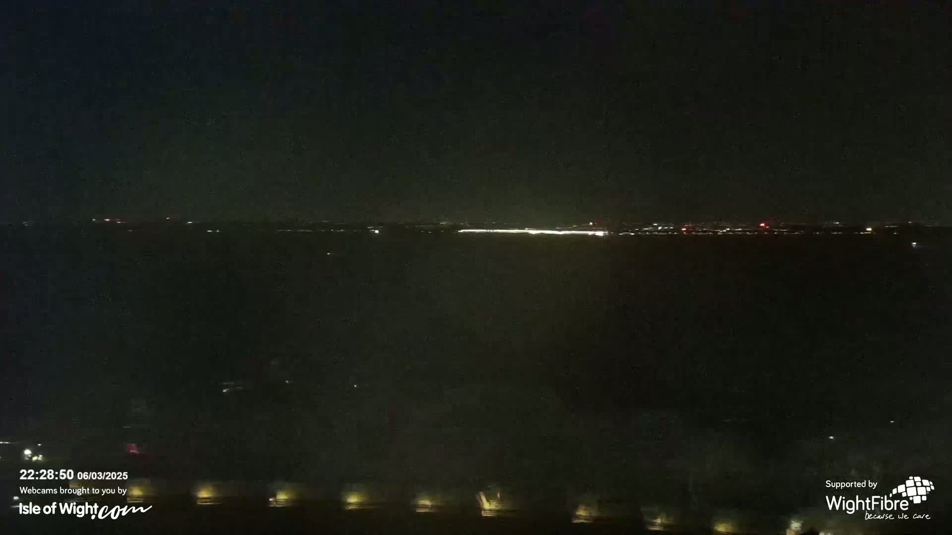 A nighttime view of a dark body of water with a distant, faintly lit cityscape on the horizon under a clear, dark sky.