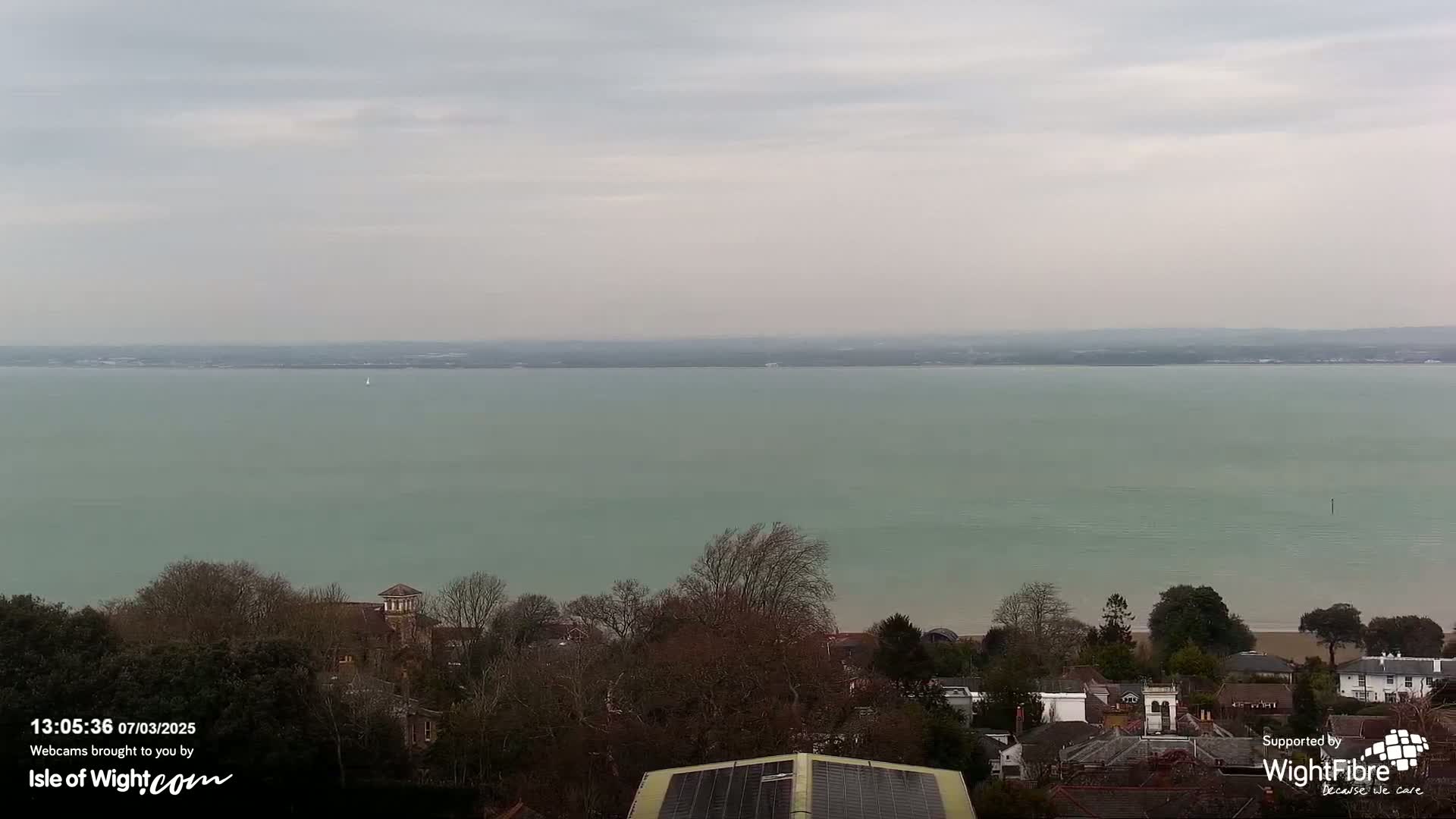 A wide, calm body of pale-green water stretches to a distant shoreline under a cloudy sky, overlooking a residential area with trees and buildings.