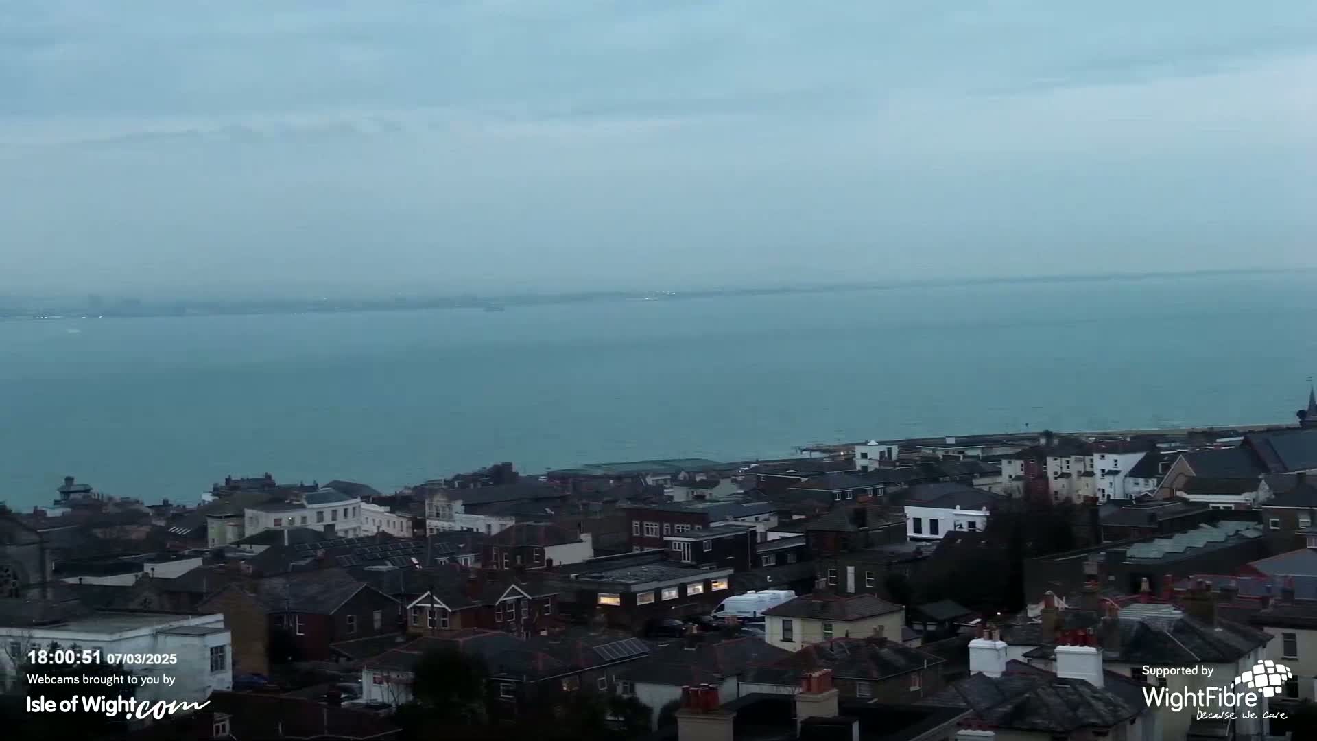 A coastal town overlooks a calm, teal sea under a hazy, overcast sky.