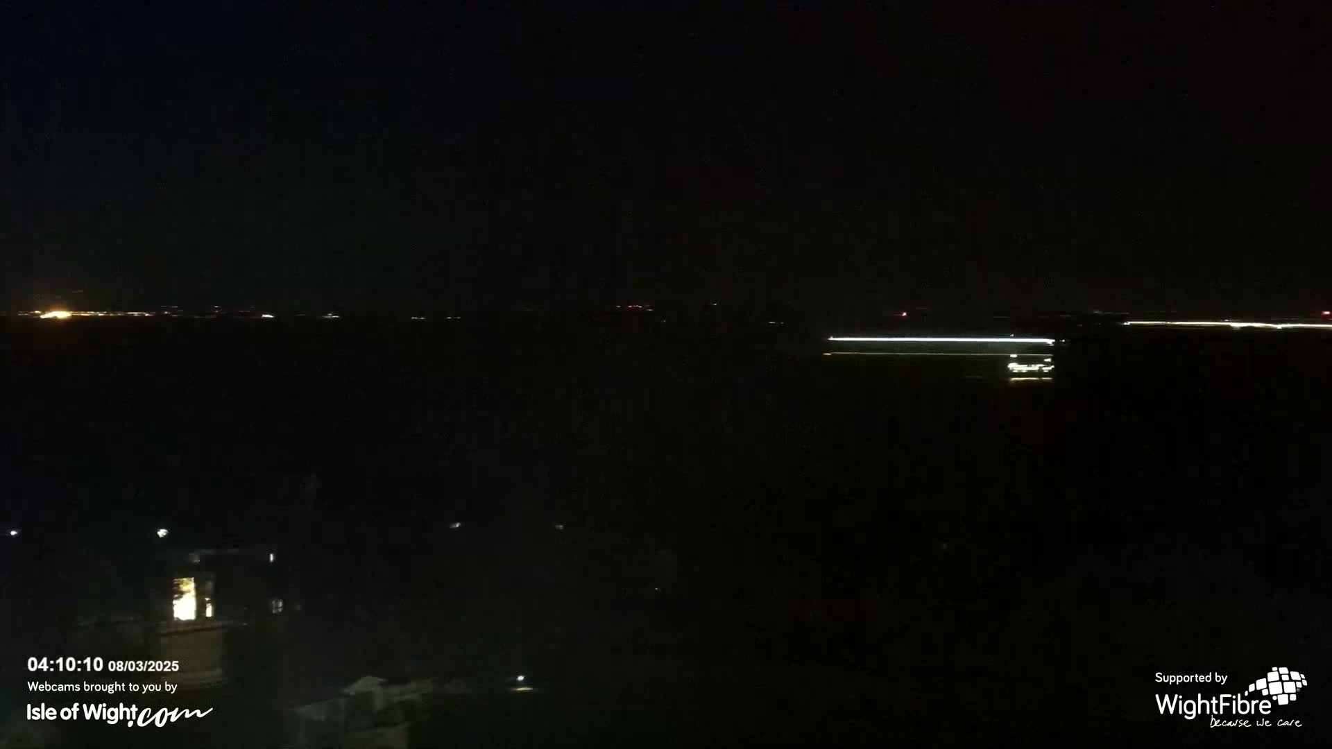 A nighttime view of a dark body of water with faint lights on the horizon and a few scattered lights on the land in the foreground.