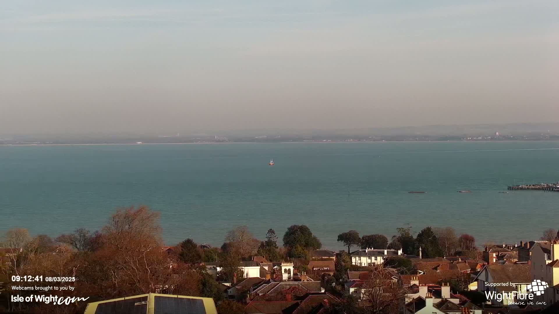 A hazy, calm sea stretches to a distant shoreline, overlooked by a residential area with brown-roofed houses and leafless trees.