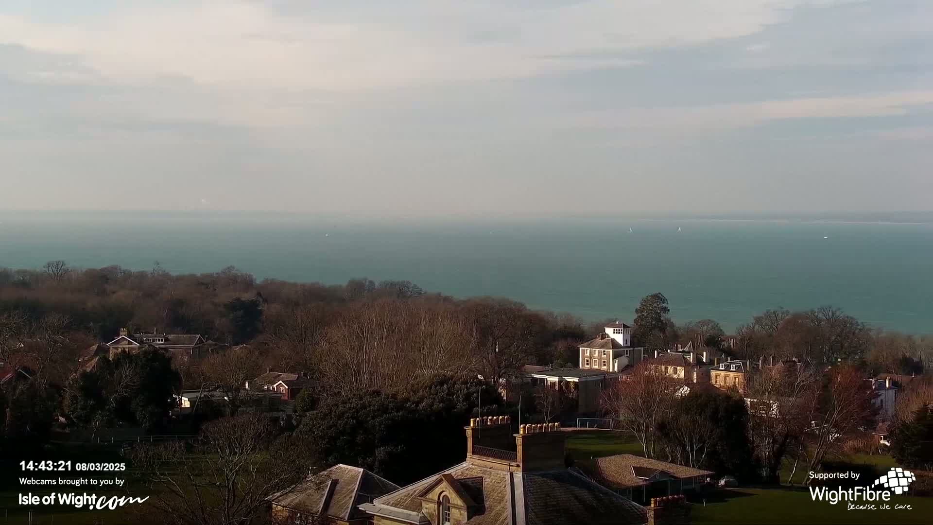 A tranquil seaside town nestled among bare winter trees overlooks a calm, teal sea under a mostly cloudy sky.