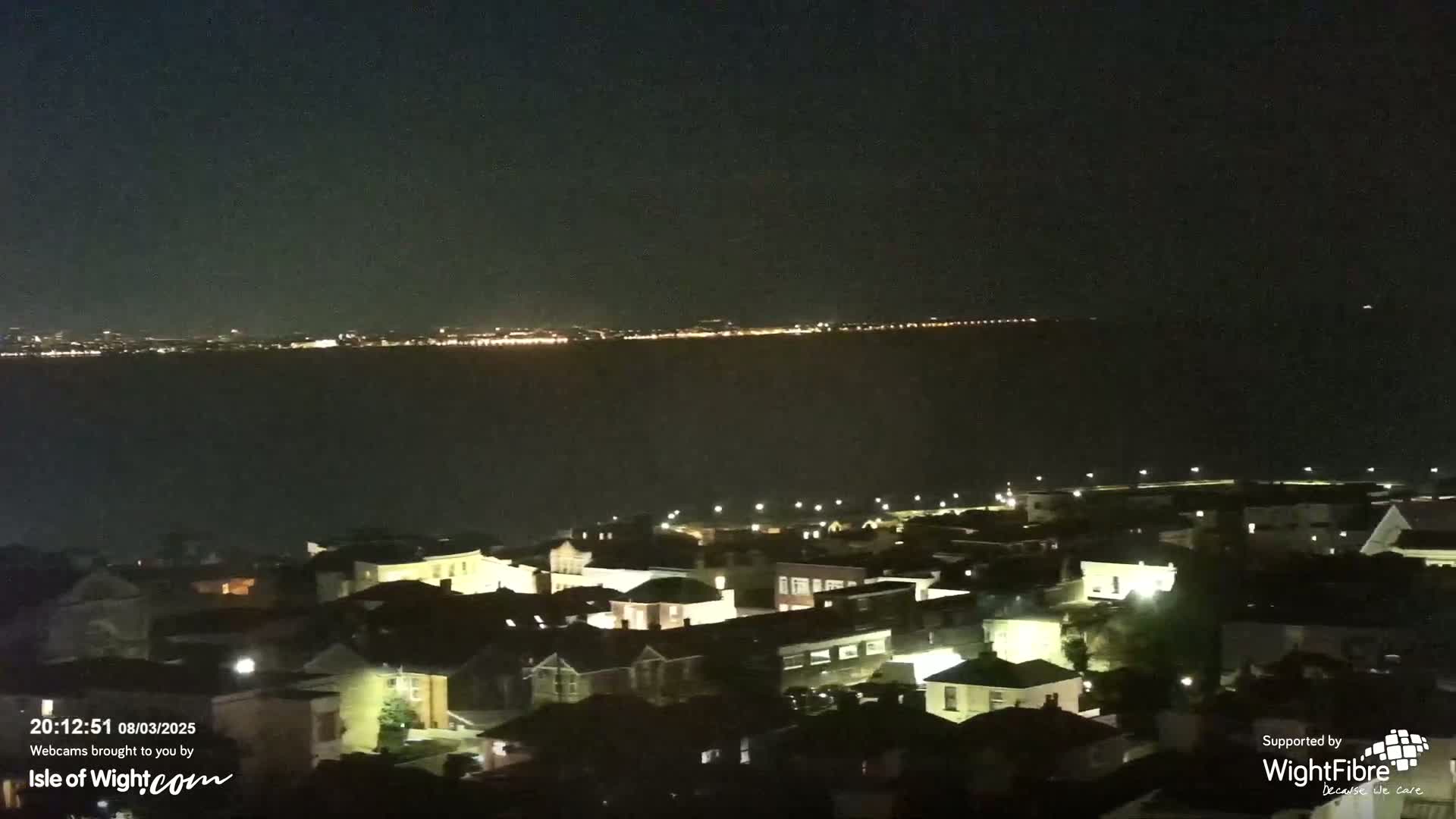 A nighttime view of a town illuminated by streetlights, overlooking a dark body of water with a distant city skyline faintly visible in the background.