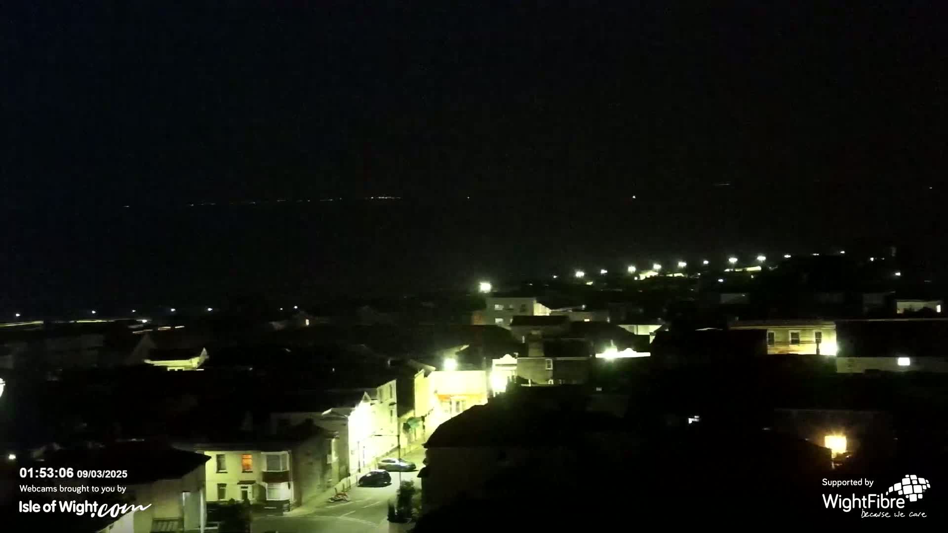 A nighttime view of a coastal town with dimly lit buildings and a dark ocean in the distance under a clear sky.