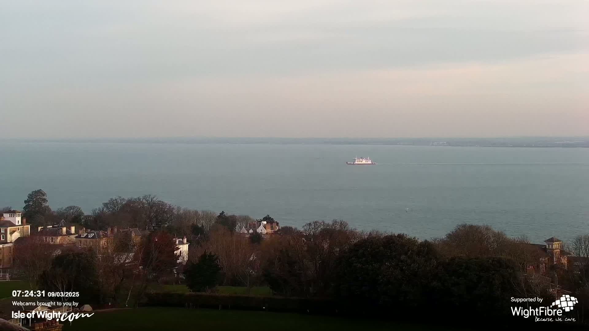 A ferry sails on a calm, gray sea, with a line of buildings and trees visible on the shore in the foreground.