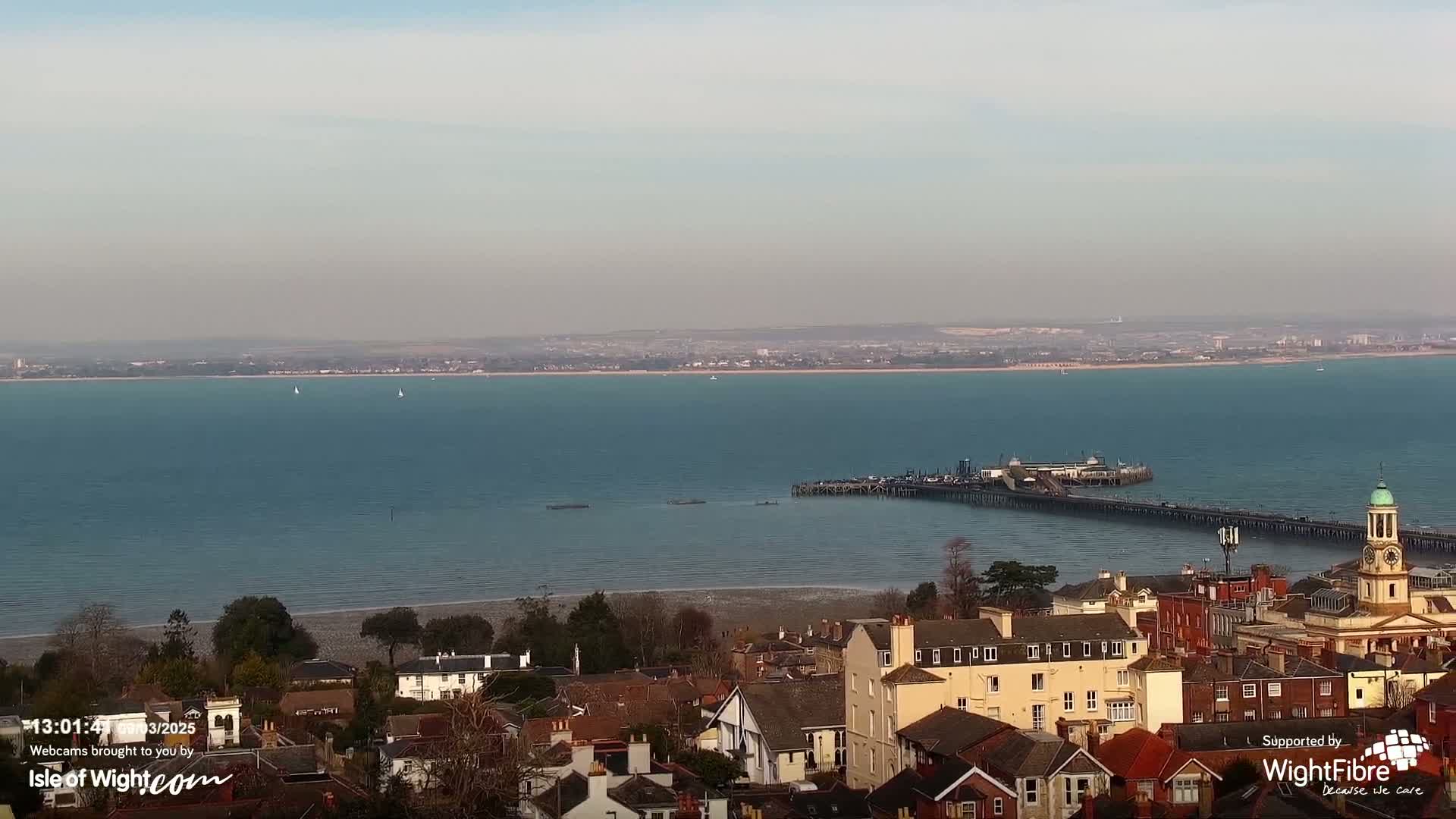 A coastal town overlooks a calm, blue bay with a long pier extending into the water under a hazy sky.