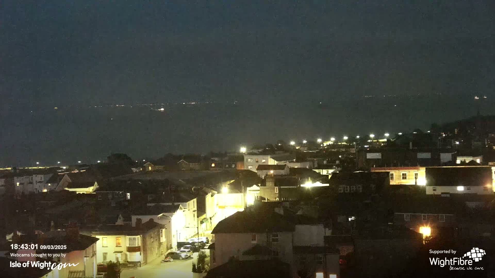 A nighttime view of a town illuminated by streetlights, overlooking a dark body of water with faint lights on the horizon under a clear, dark sky.