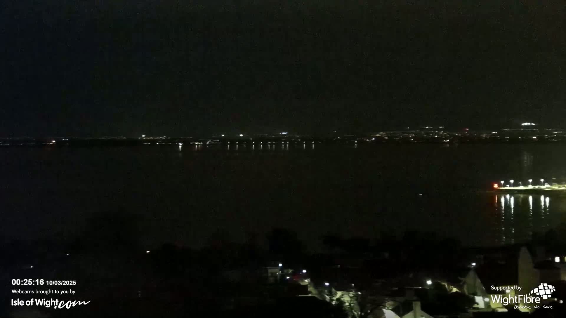 A nighttime view of a dark body of water reflecting city lights on the far shore, with a few lights visible closer in the foreground.