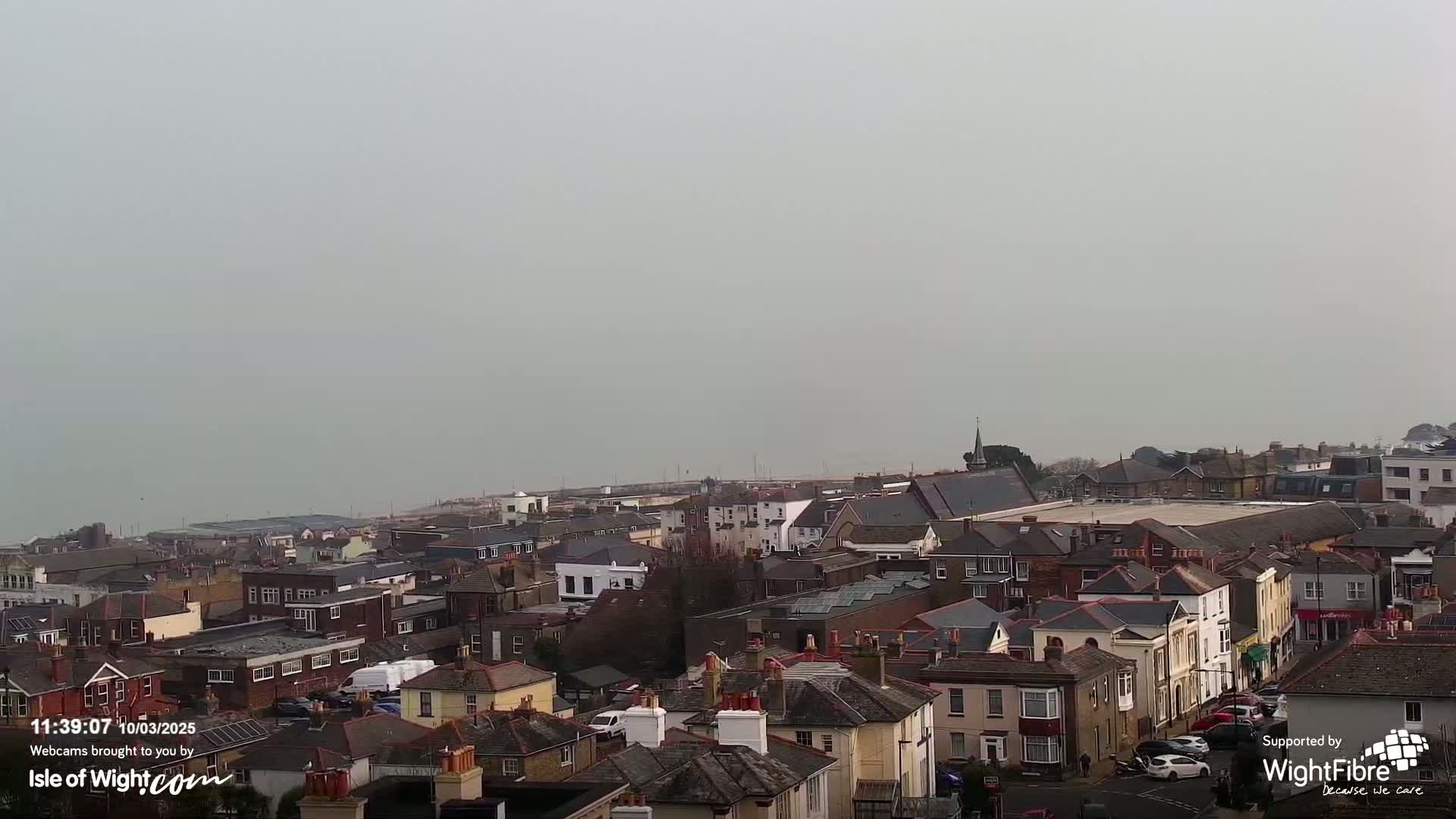 A hazy, overcast day reveals a coastal town with densely packed houses and buildings, extending to a calm sea in the distance.