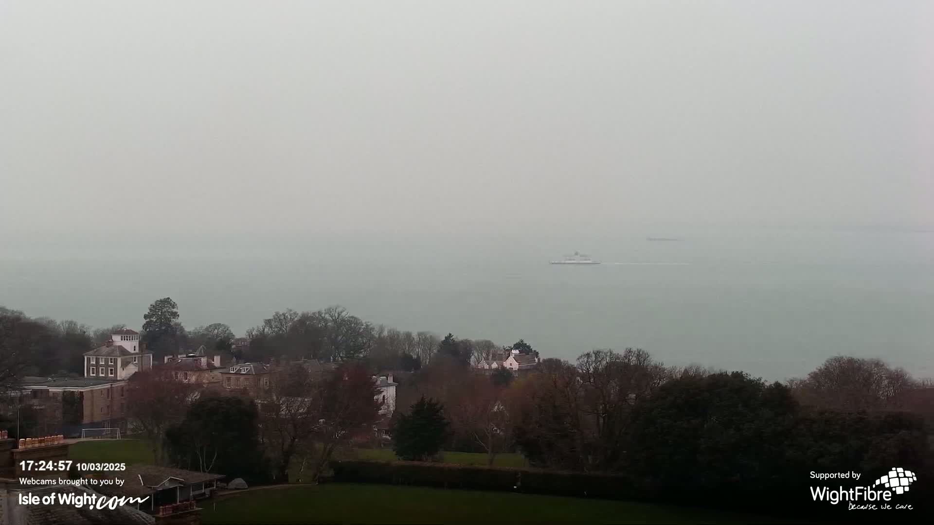 A hazy, overcast view shows a ferry on a calm body of water, with bare winter trees and buildings visible in the foreground.