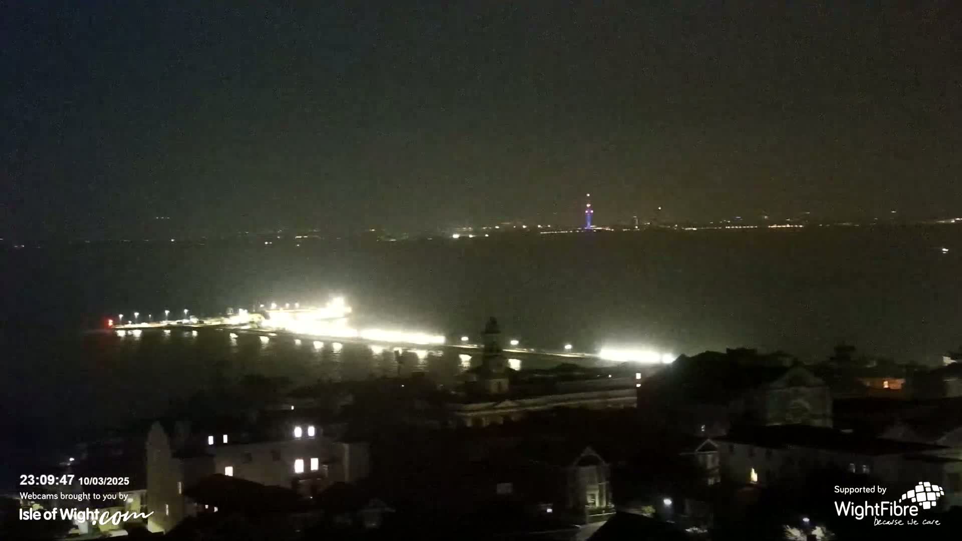 A nighttime view of a city waterfront, with a body of water reflecting lights from buildings and a distant shoreline.