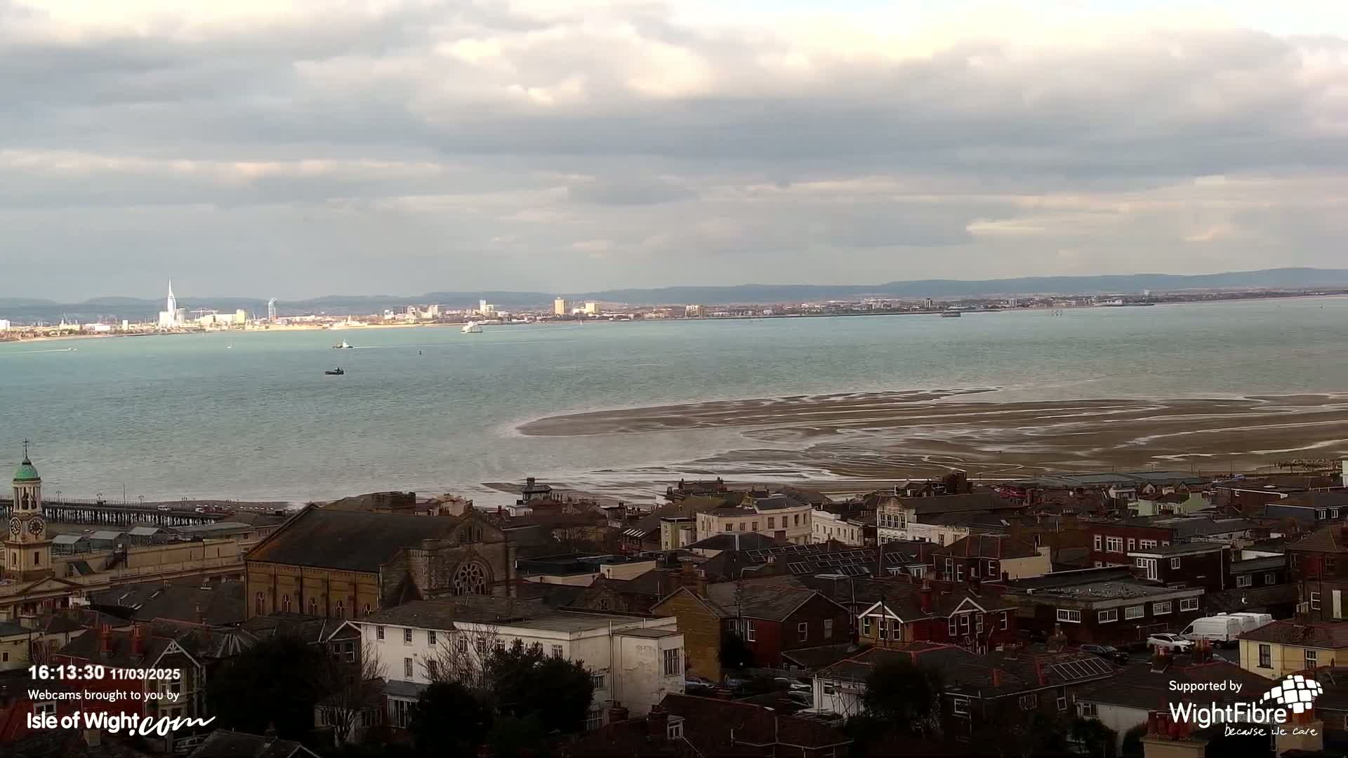 A coastal town with a church and many houses overlooks a large body of water with exposed sandbars under a cloudy sky.