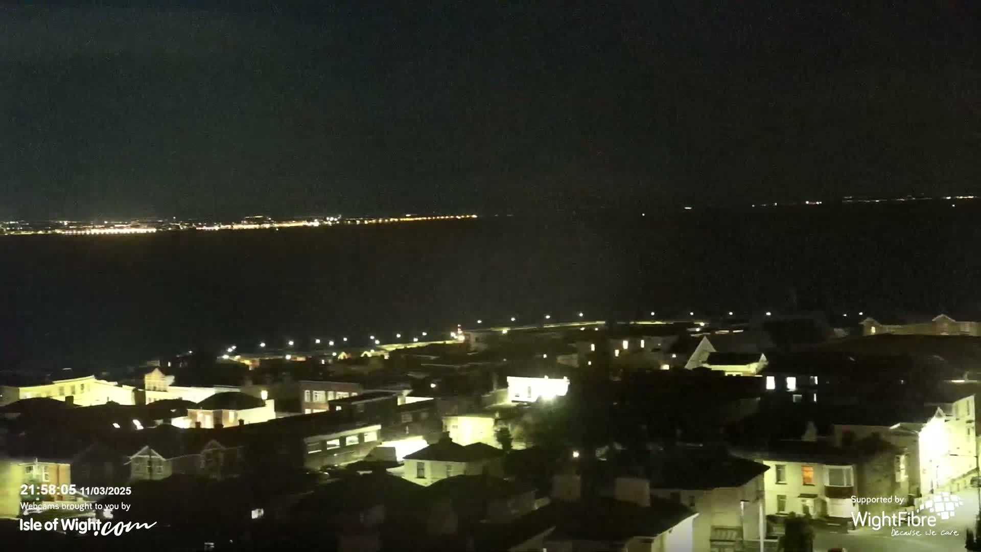 A nighttime aerial view shows a coastal town with lit houses and buildings, overlooking a dark body of water with a distant city skyline visible on the horizon under a clear, dark sky.