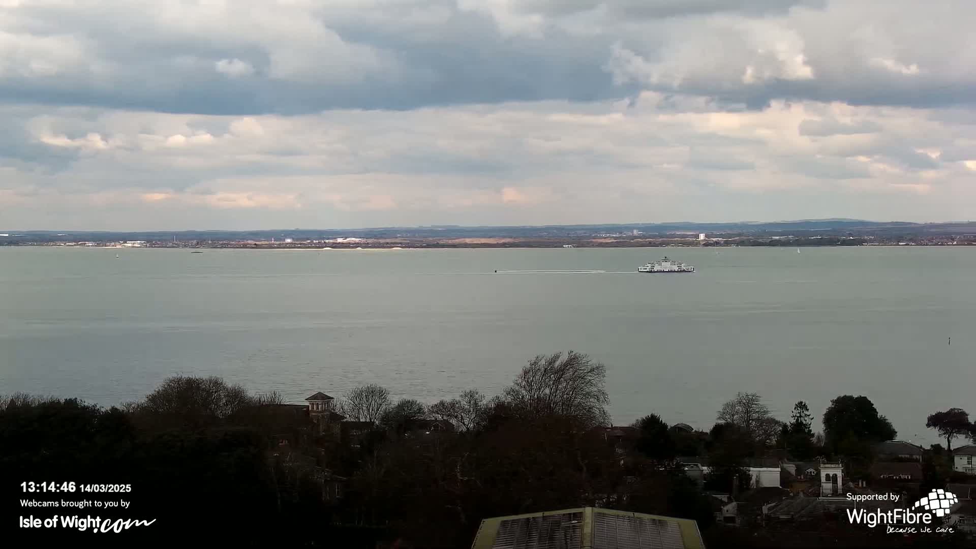 A ferry travels across a calm, gray body of water under a mostly cloudy sky, with a distant shoreline and tree-lined residential area visible in the foreground.