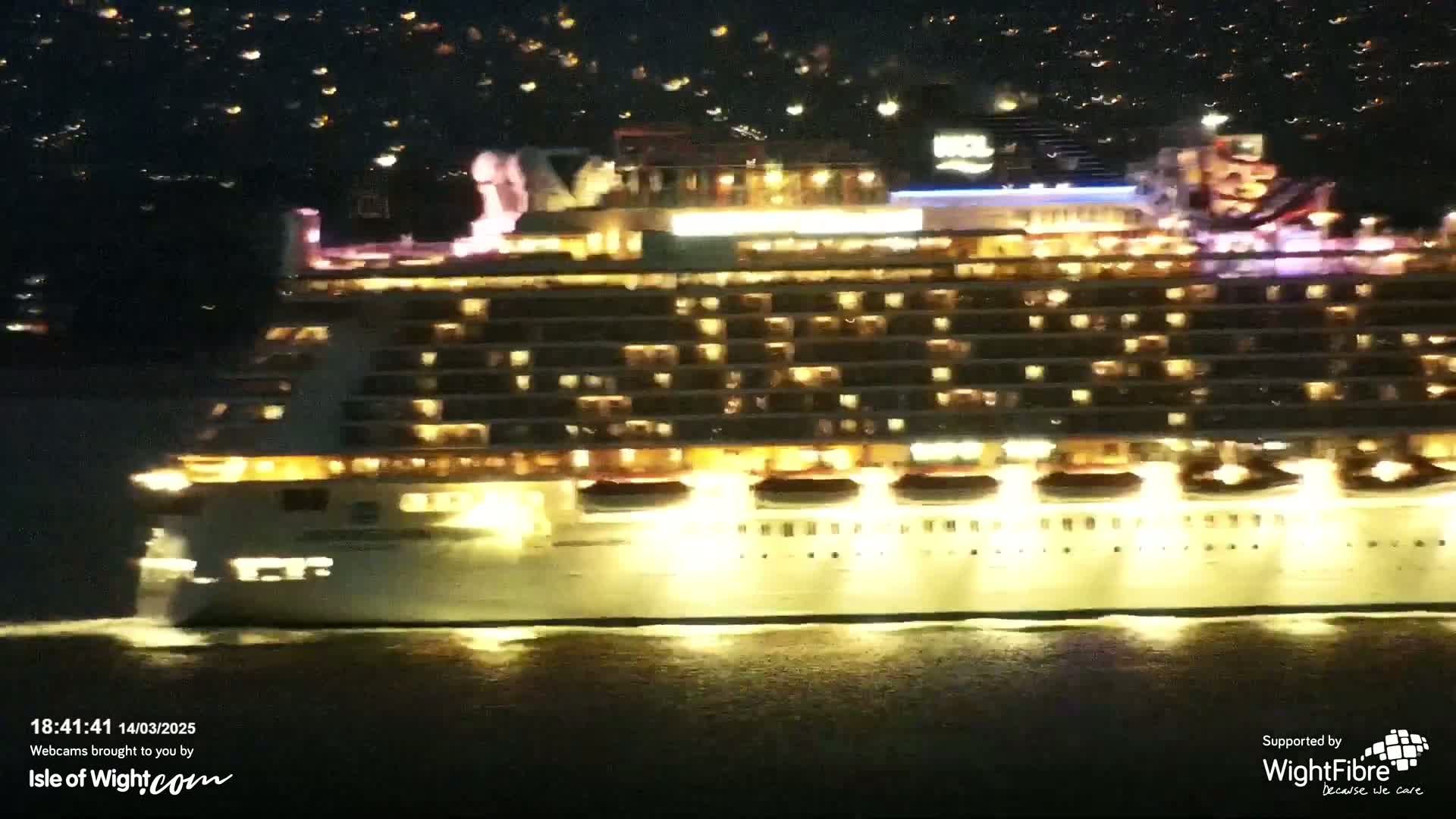 A large, brightly lit cruise ship sails on dark water at night, with a city's lights visible in the distance.