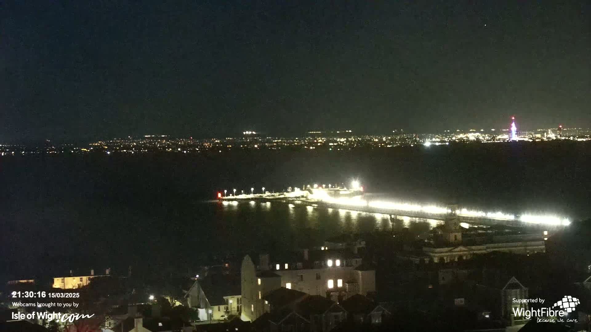 A brightly lit pier extends into a dark body of water at night, with a city skyline visible in the distance under a clear sky.