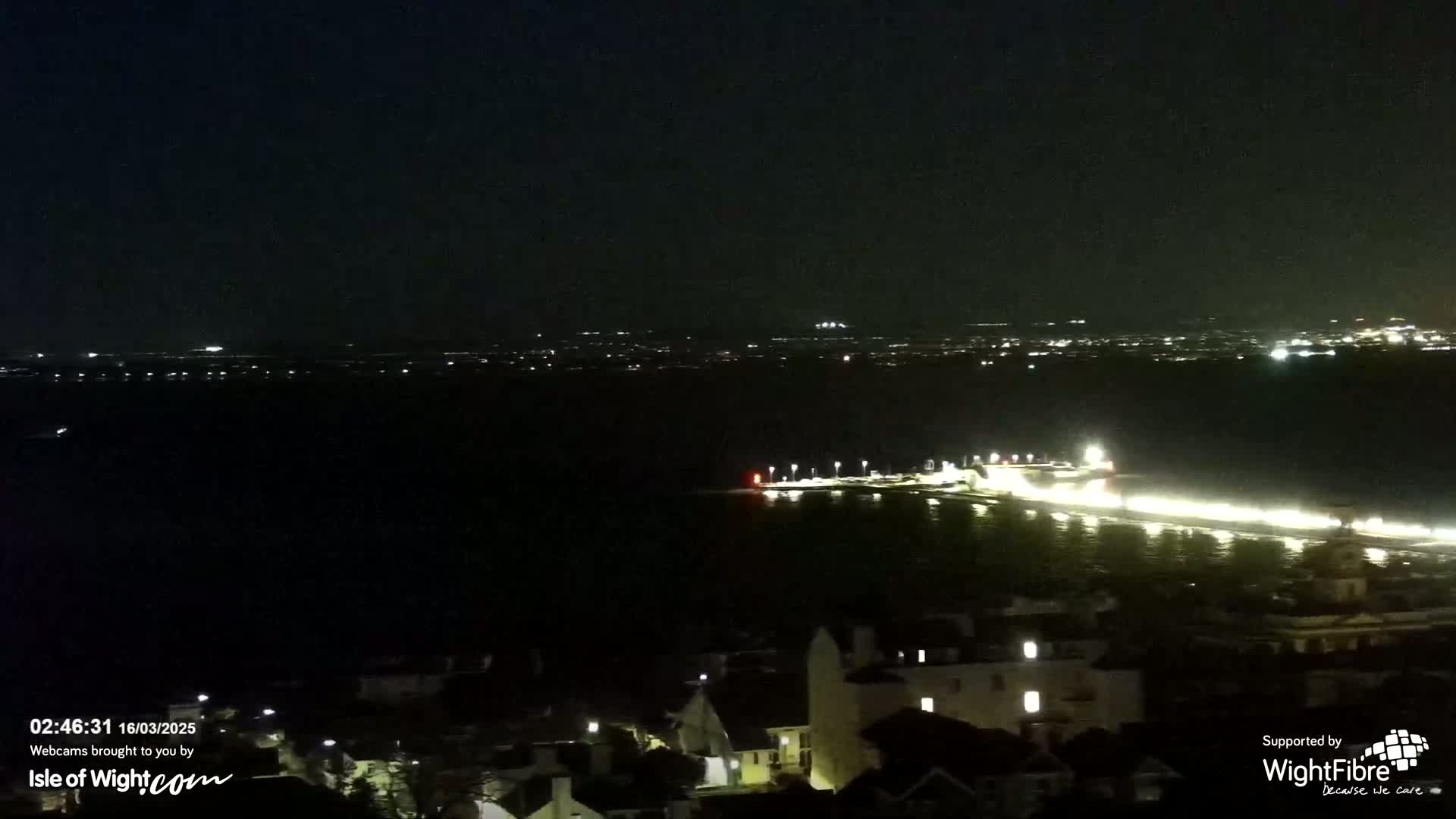 A nighttime view of a dark body of water with a lit-up shoreline and distant city lights under a clear sky.