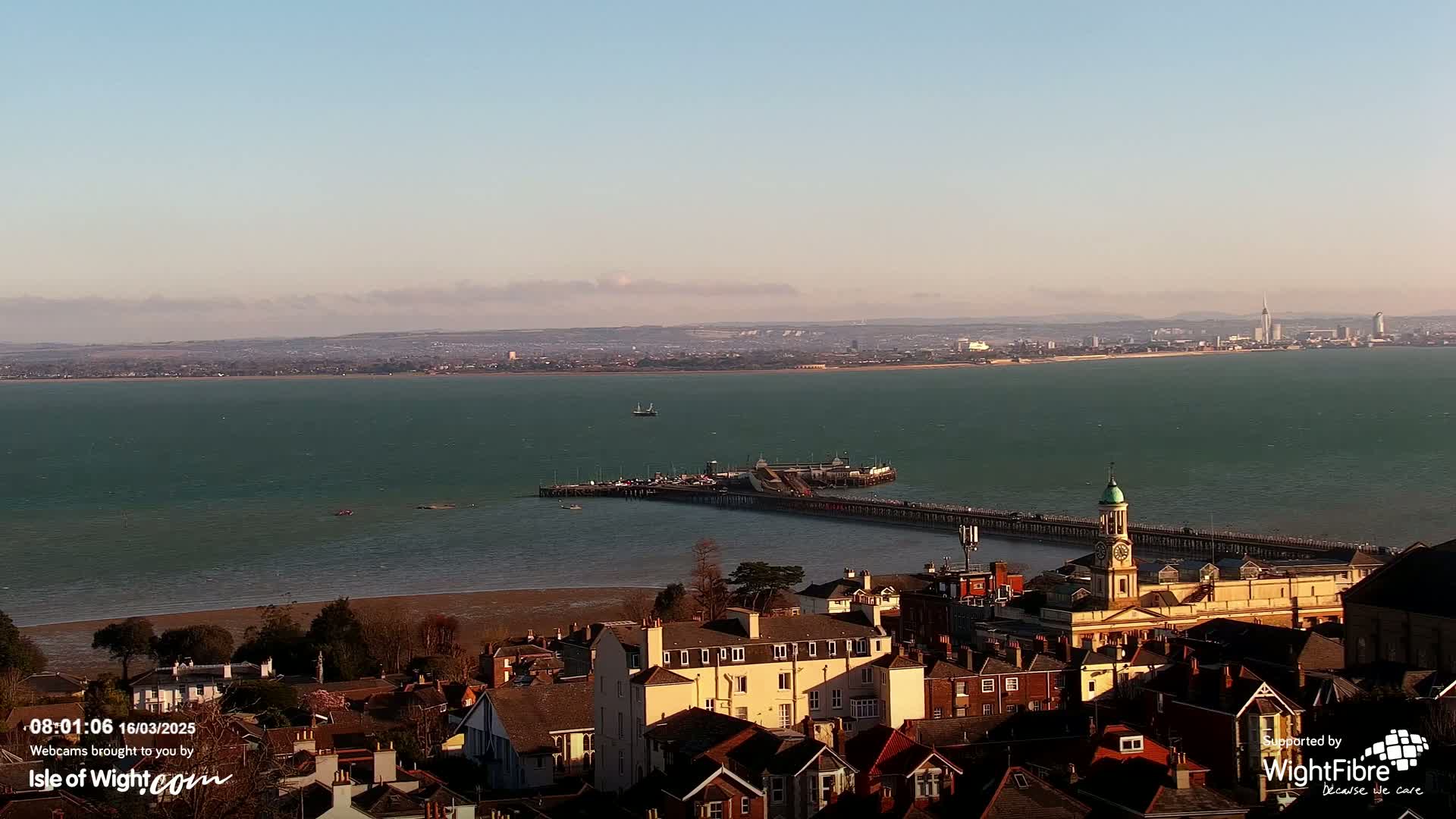 A coastal town with a long pier extends into a teal sea under a mostly clear sky, with a distant city skyline visible on the horizon.