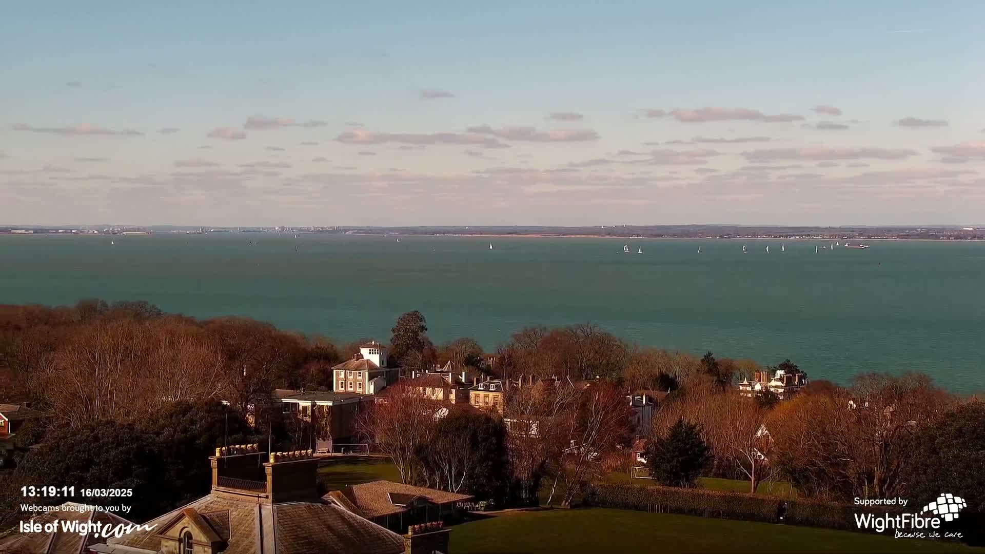 An aerial view shows a teal expanse of water with sailboats, under a partly cloudy sky, overlooking a collection of buildings nestled among leafless trees.