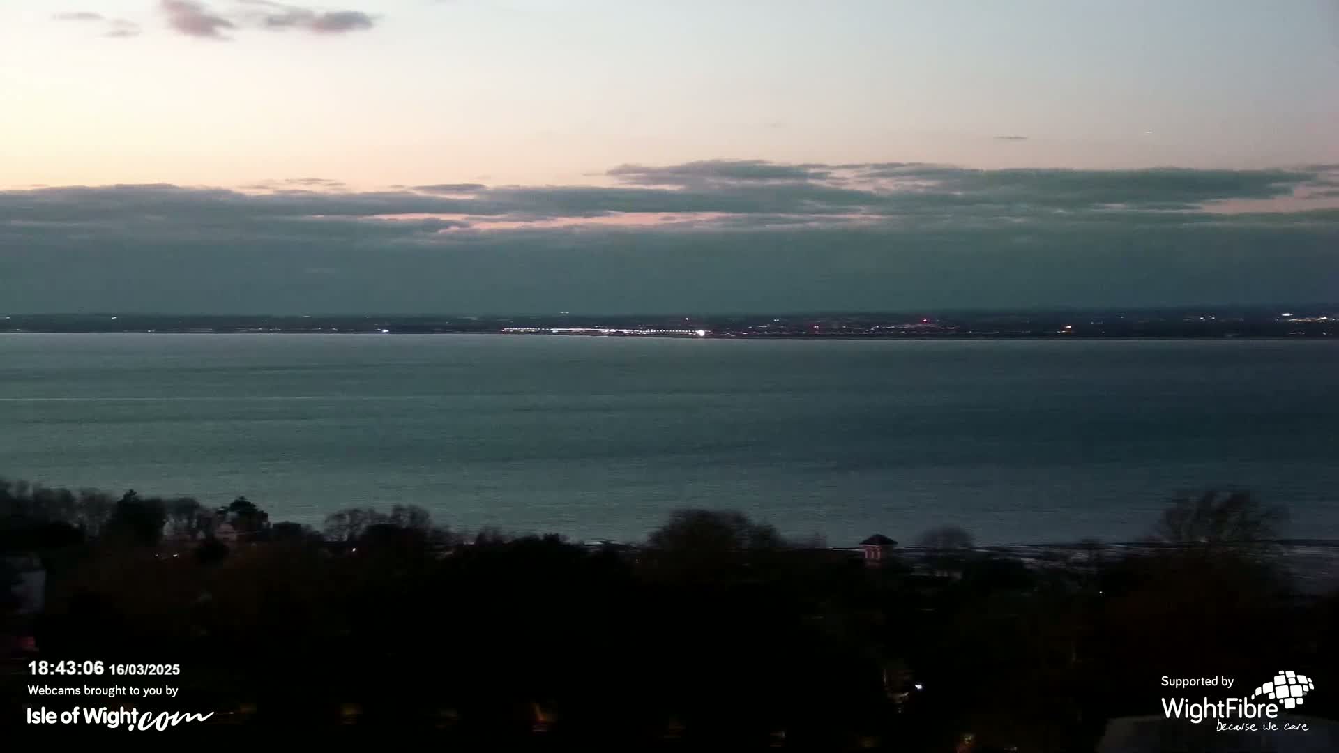 A wide shot of a calm body of water at dusk under a cloudy sky, with a dark shoreline and distant lights visible across the water.