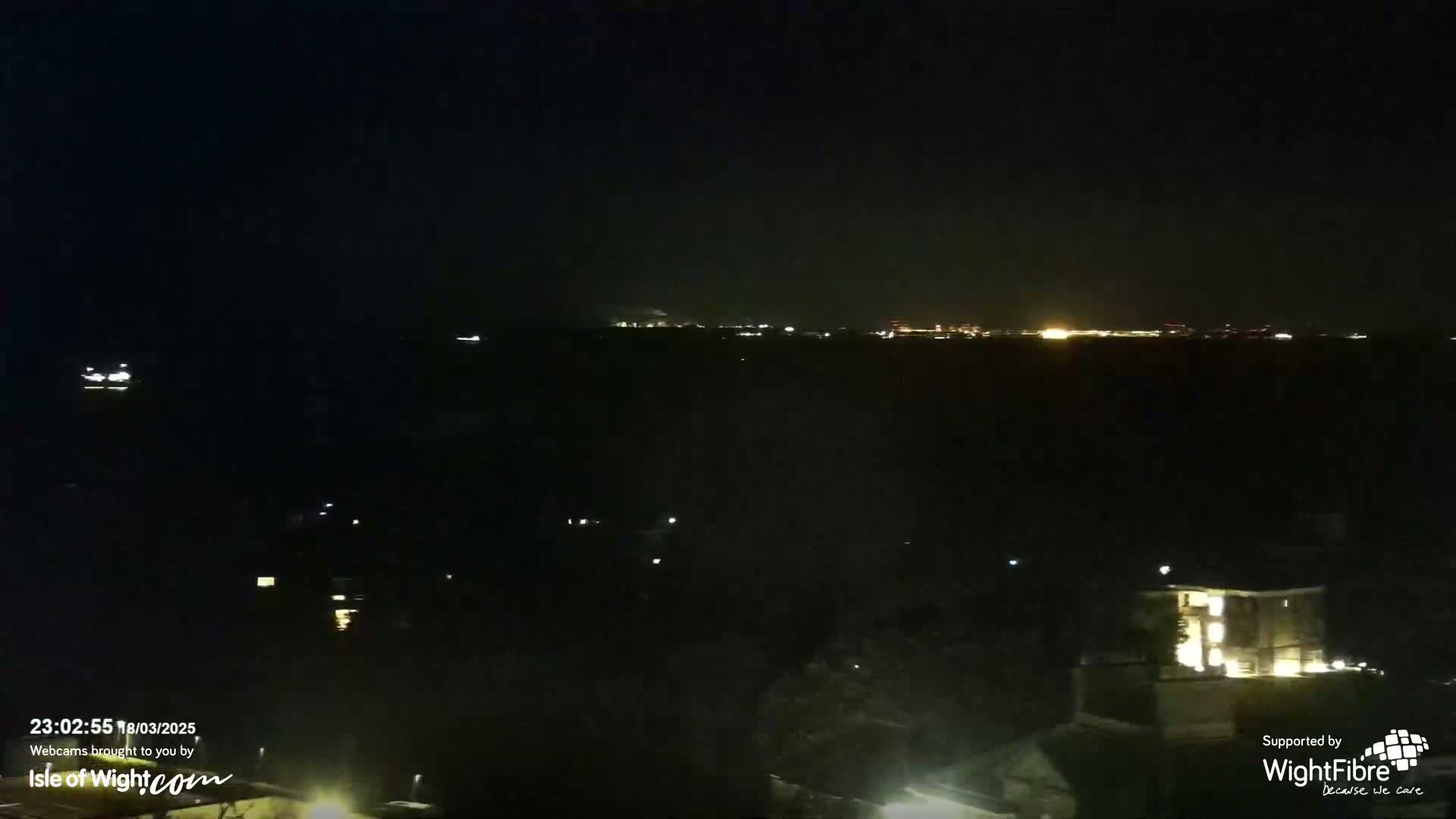A nighttime long shot shows a dark body of water with a distant city skyline faintly illuminated, and some lights visible on the shoreline in the foreground.