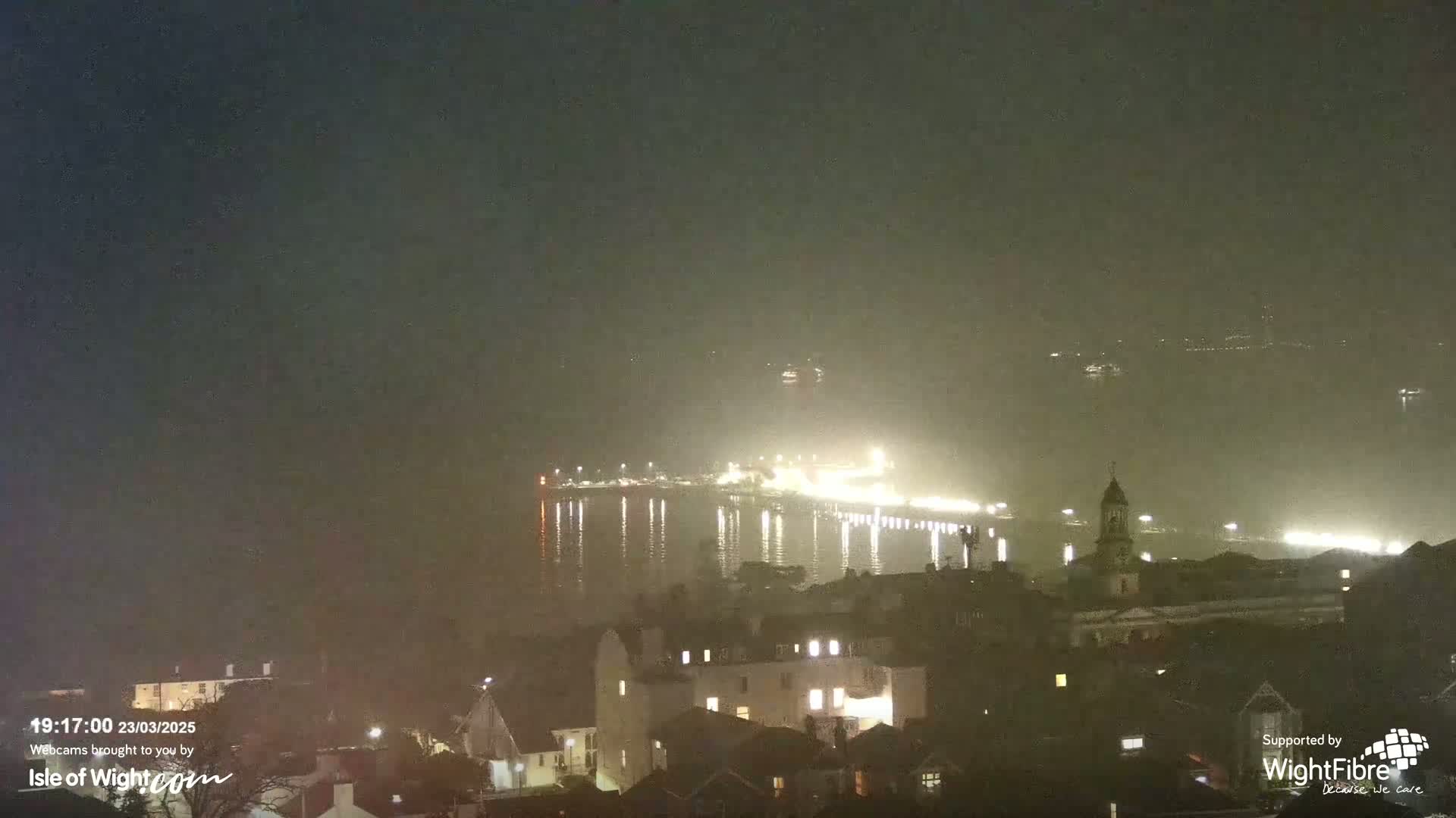 A brightly lit pier extends into a foggy, dark night over calm water, with a town visible in the foreground.