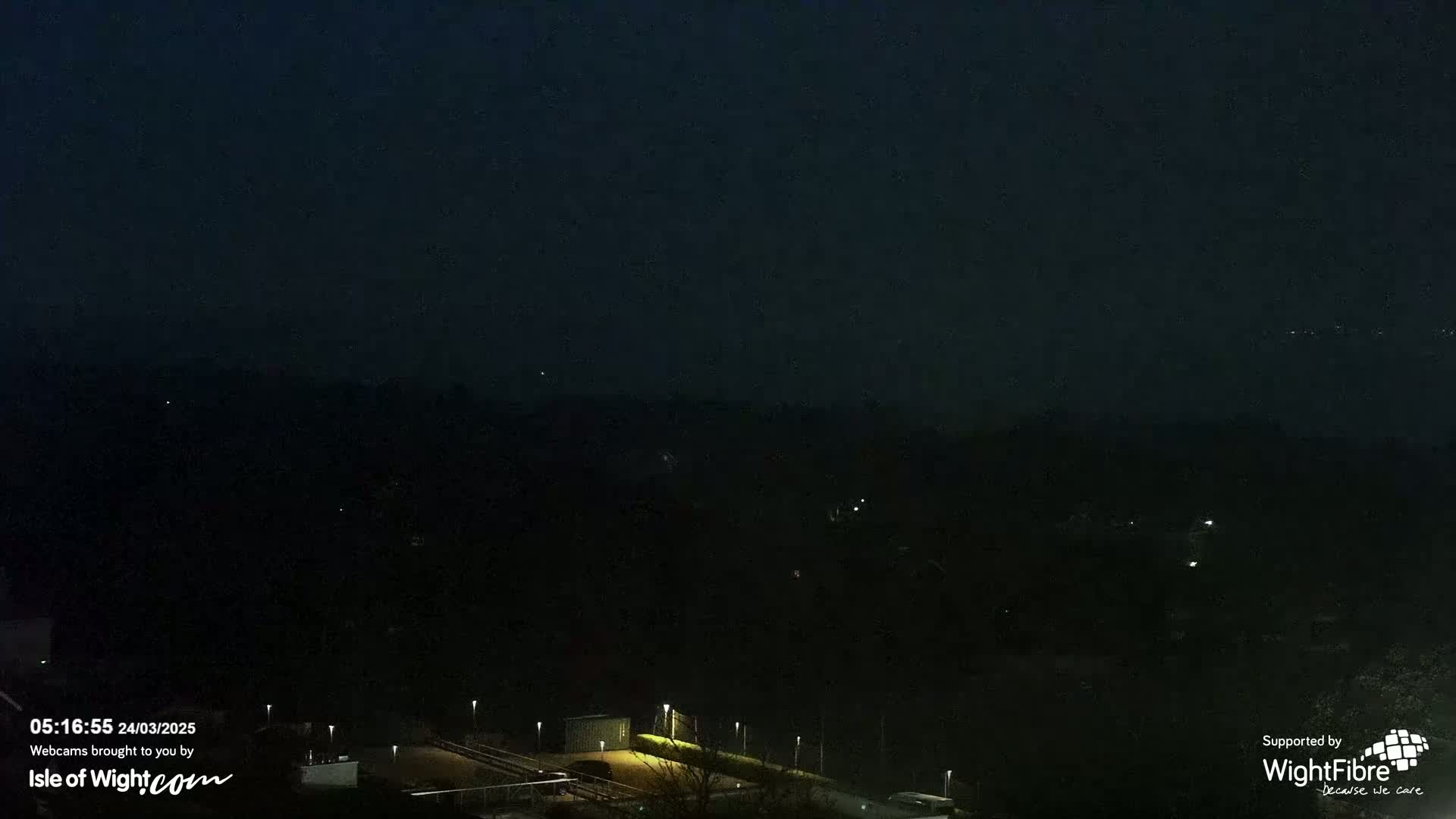 A dark, nighttime view of a landscape with sparsely lit buildings and trees under a clear, dark sky.