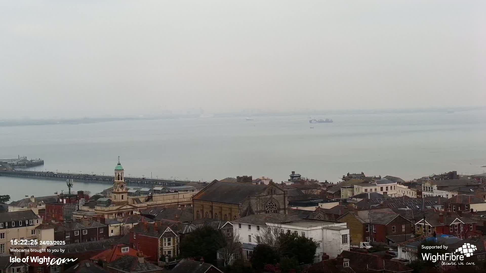 A hazy, overcast view shows a town with a church and pier overlooking a calm body of water.