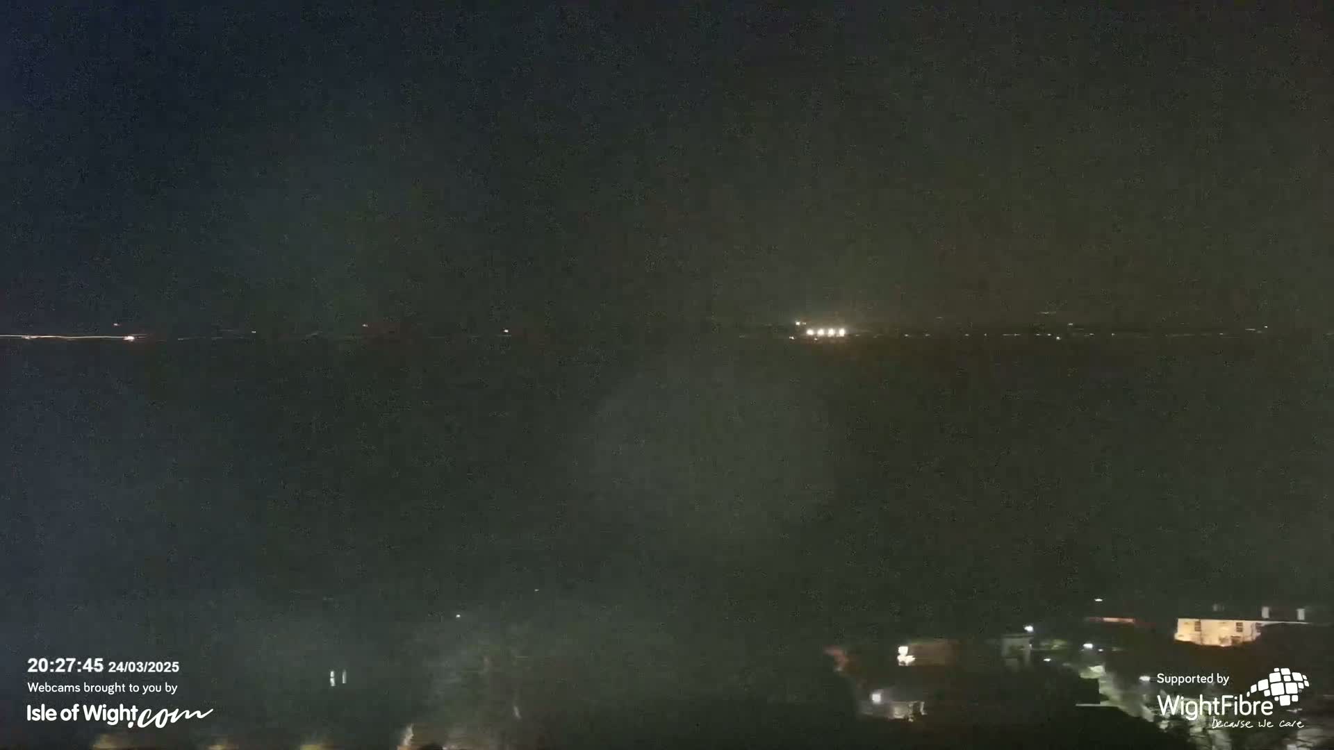 A nighttime long shot shows a dark, possibly hazy or foggy, body of water with faint lights on the far shore and some dimly lit buildings closer to the viewer.