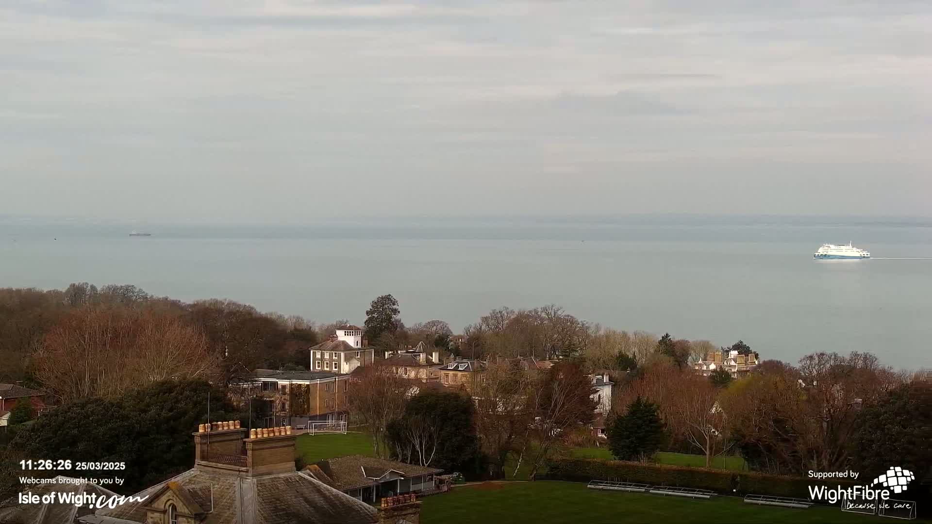 A ferry is visible on a calm, gray sea, beyond a collection of buildings and bare winter trees on a grassy hillside.