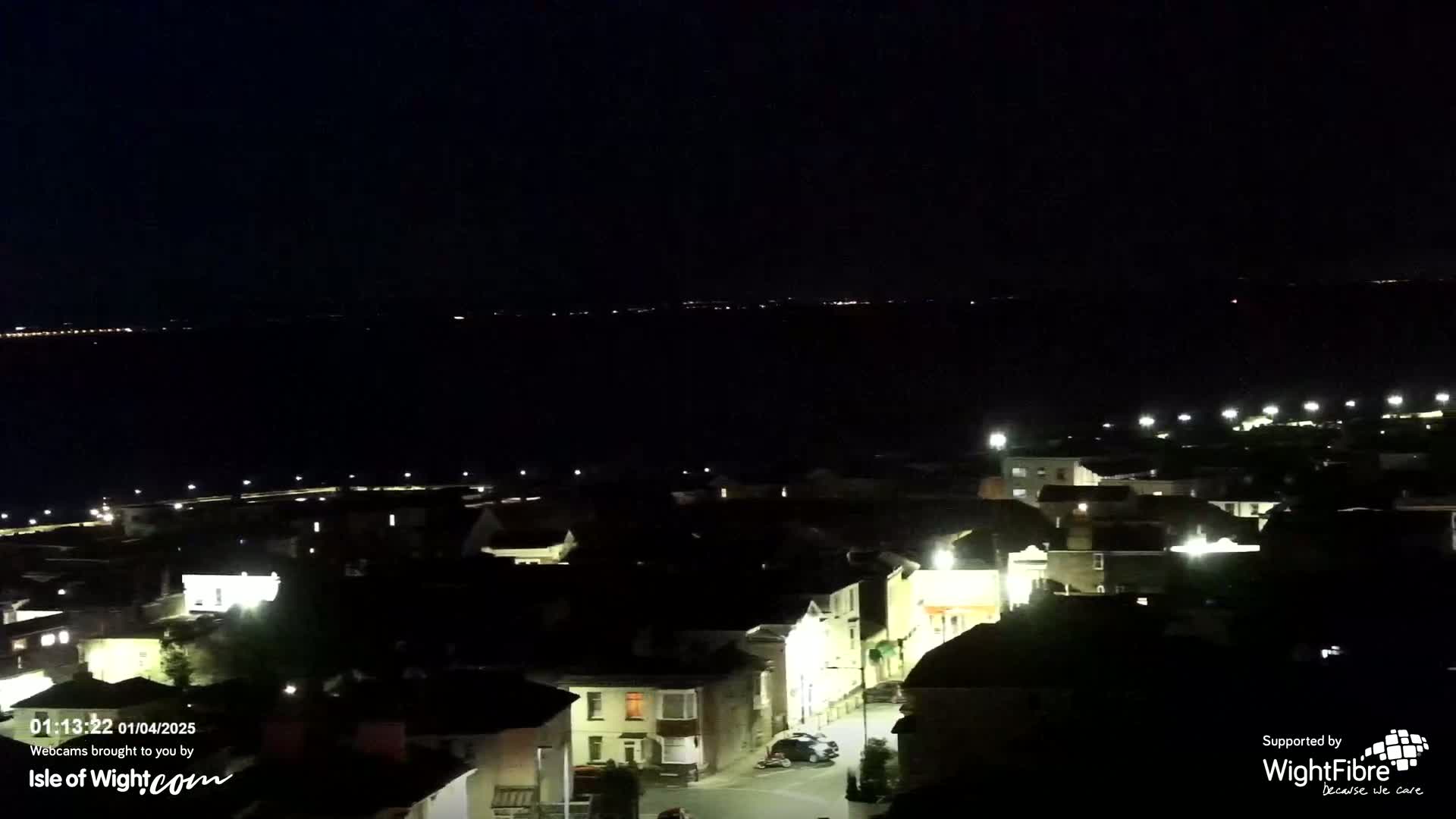 A nighttime, high-angle view shows a town's illuminated streets and buildings next to a dark body of water with faintly visible lights on the far shore.