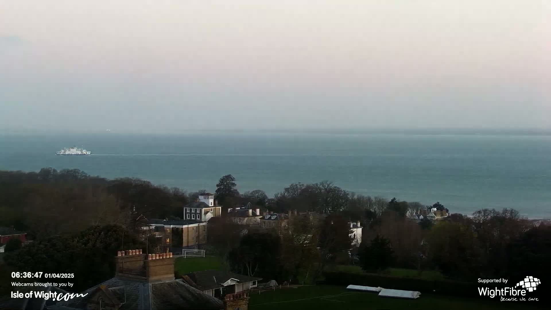 A ferry sails on a calm, hazy sea past a collection of buildings nestled among trees on a hill.
