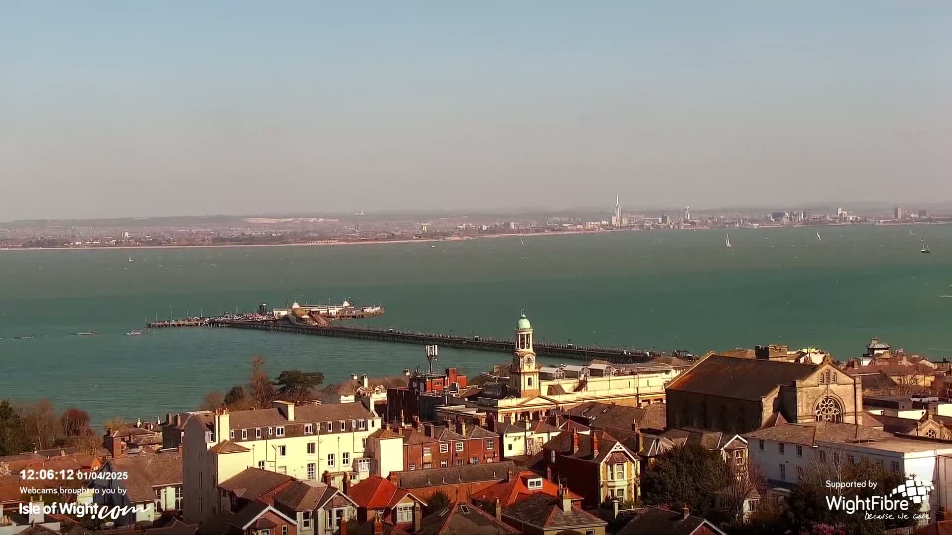 A coastal town with a long pier extending into a teal sea under a sunny sky, with a distant cityscape visible across the water.