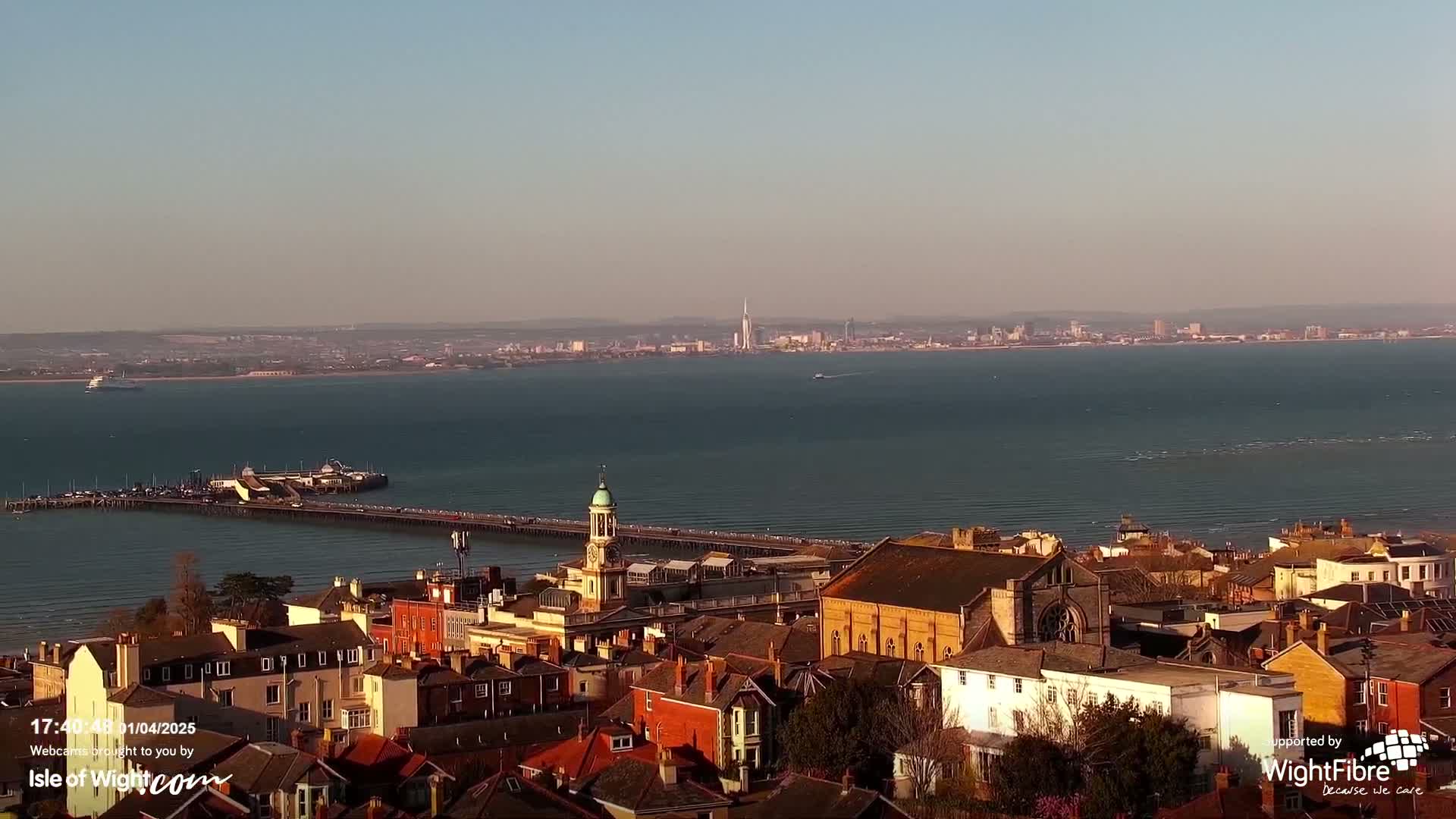 A coastal town overlooks a calm body of water with a distant city skyline visible under a clear sky.