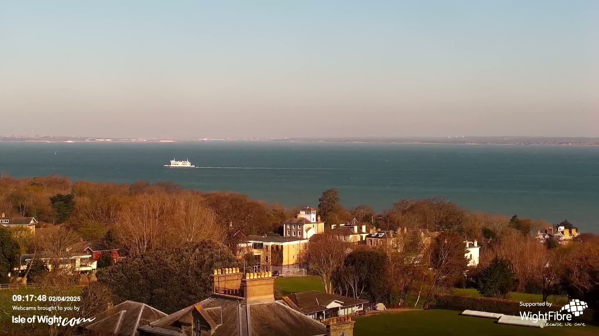 A ferry travels across a calm body of water under a clear, pale blue sky, beyond a collection of houses and trees nestled on a hillside.