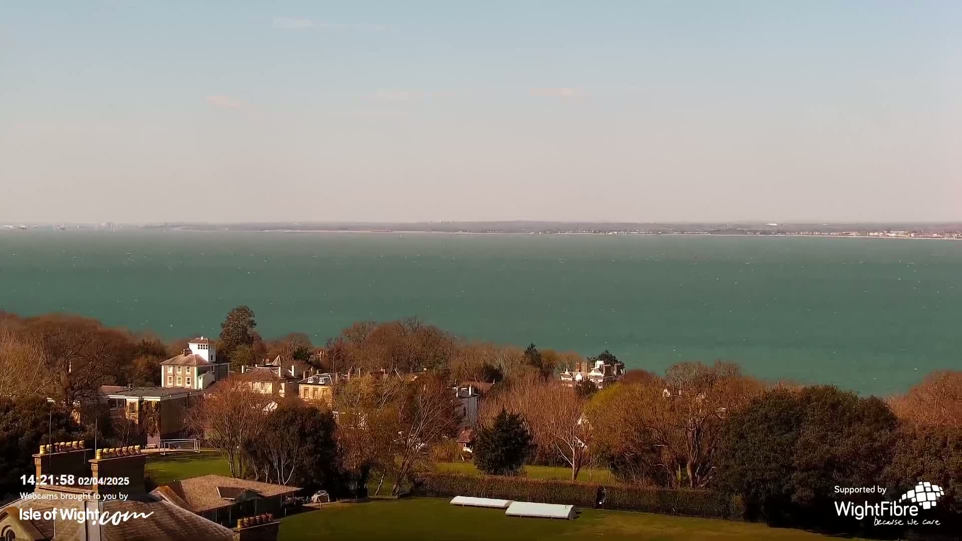A large body of teal water stretches to a distant shoreline under a clear sky, overlooking a collection of buildings nestled among bare trees on a grassy hill.