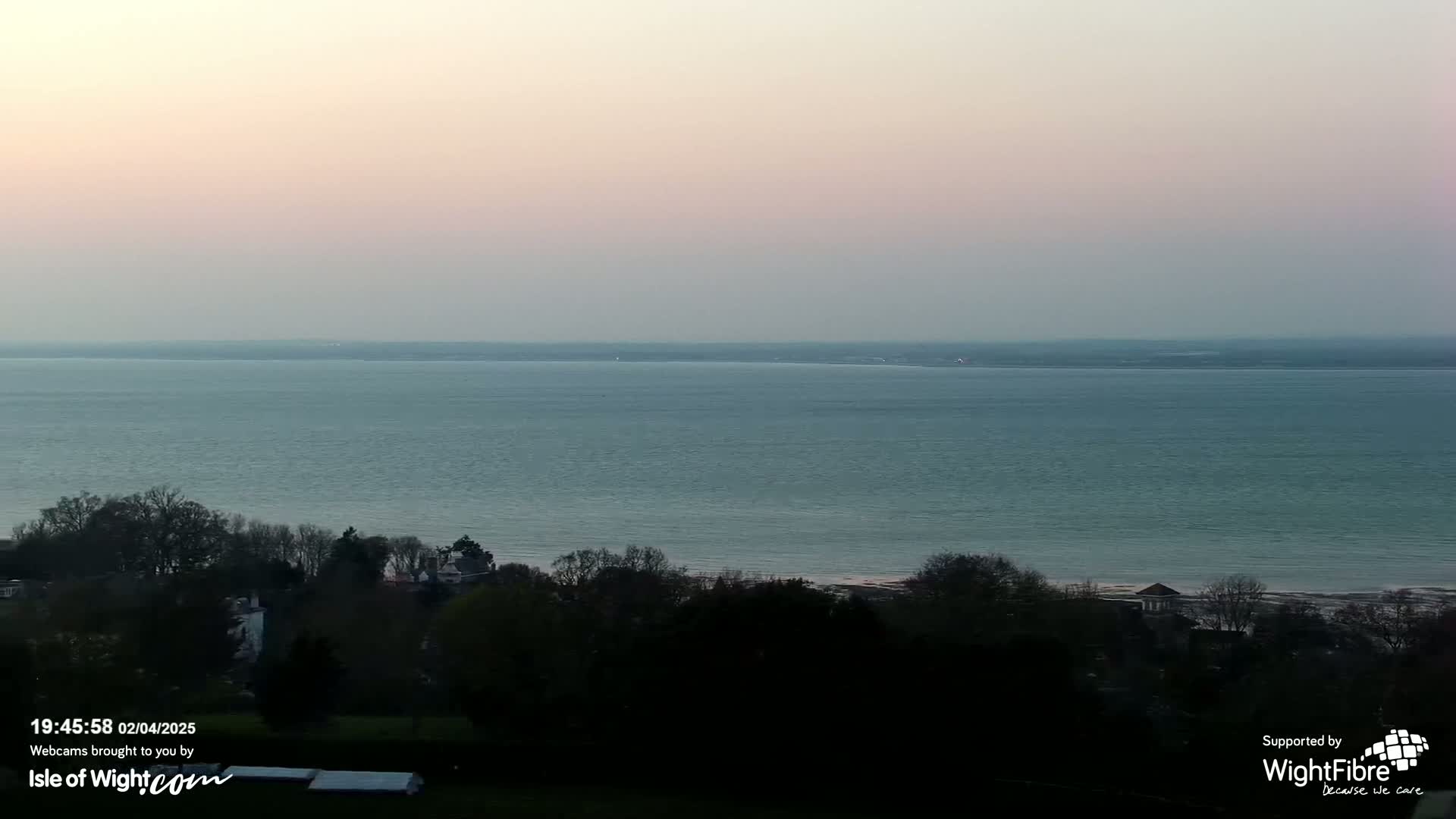 A tranquil, pale-pink sunset illuminates a calm expanse of sea, viewed from a verdant hillside dotted with dark trees.