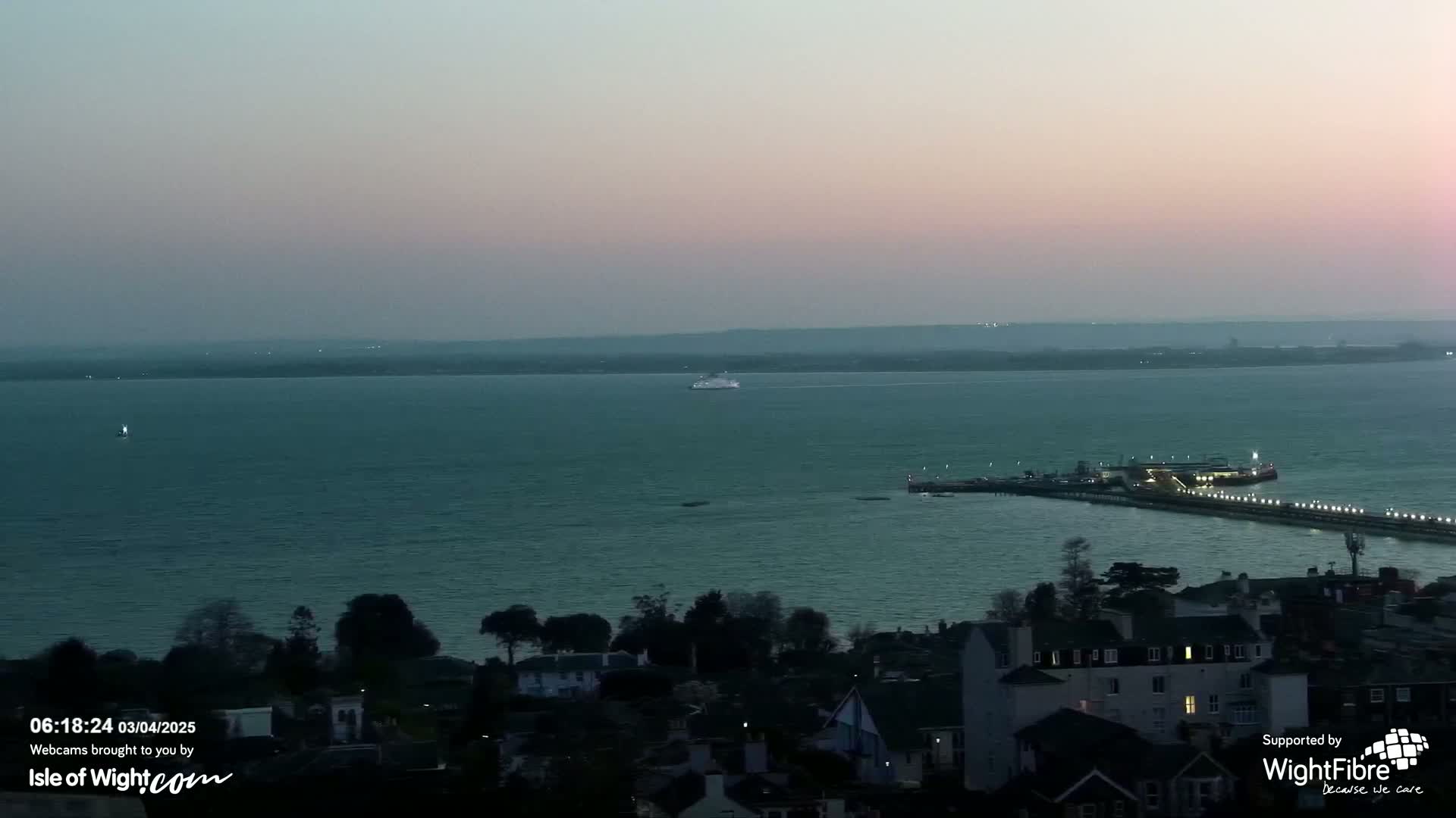 A ferry sails across a calm, dark-teal body of water at dusk, past a lit pier and a town with houses.