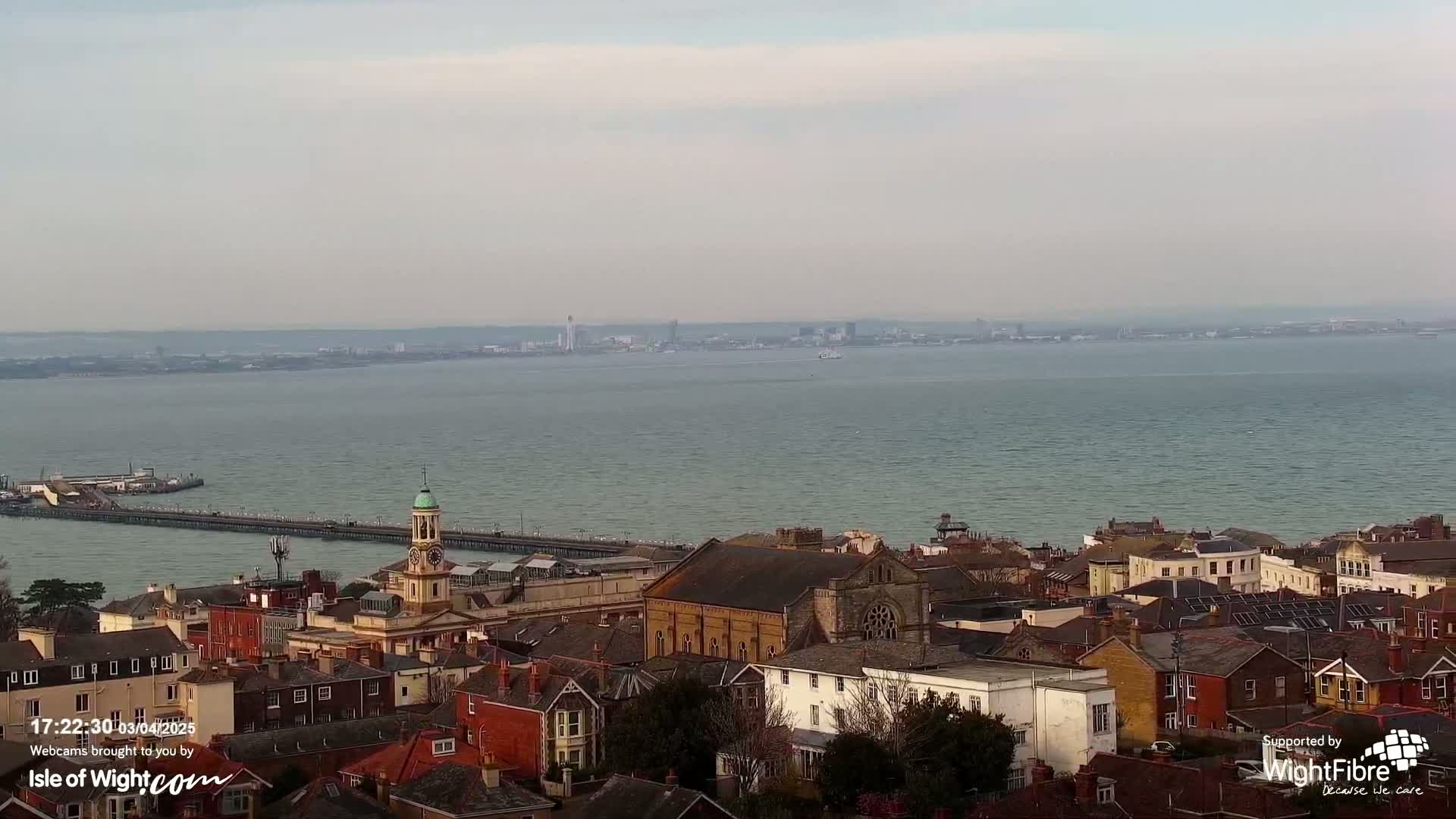An aerial view shows a town with a clock tower and pier overlooking a calm body of water with a distant cityscape under a cloudy sky.