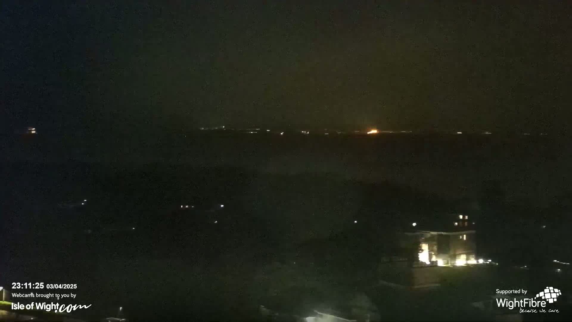 A nighttime view shows a dark body of water with a few distant lights, and a dark landmass with a few illuminated buildings closer to the viewer.