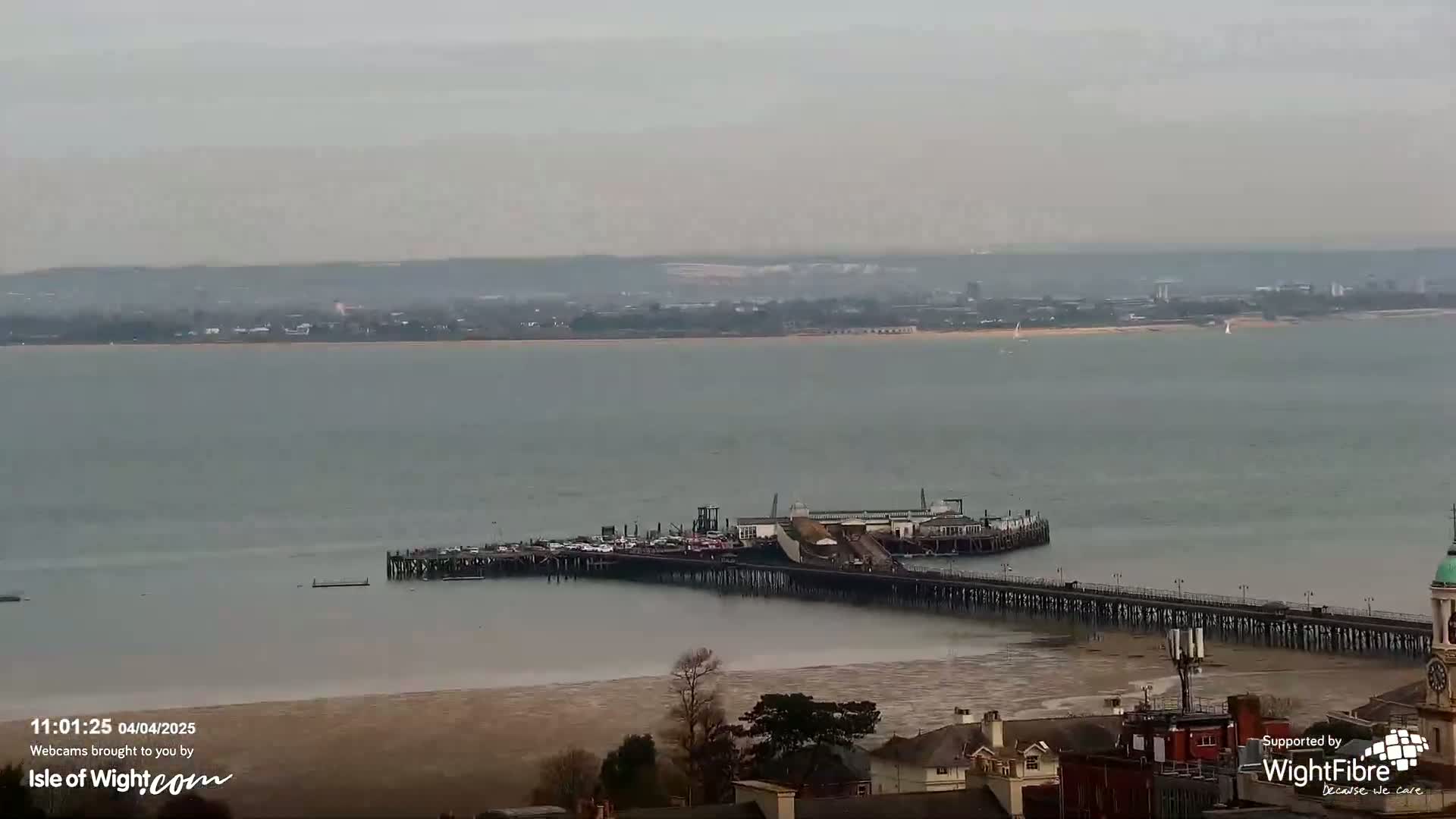 A long pier extends into a calm, gray body of water under an overcast sky, with a distant shoreline visible in the background.