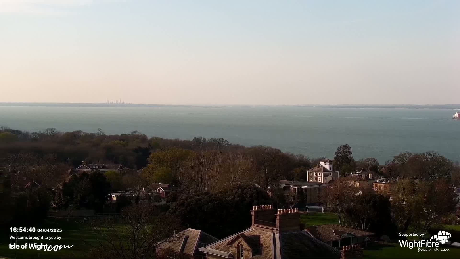 A tranquil coastal scene shows a calm body of water with a distant industrial area under a clear, pale sky, overlooking a tree-lined hillside with houses and buildings.