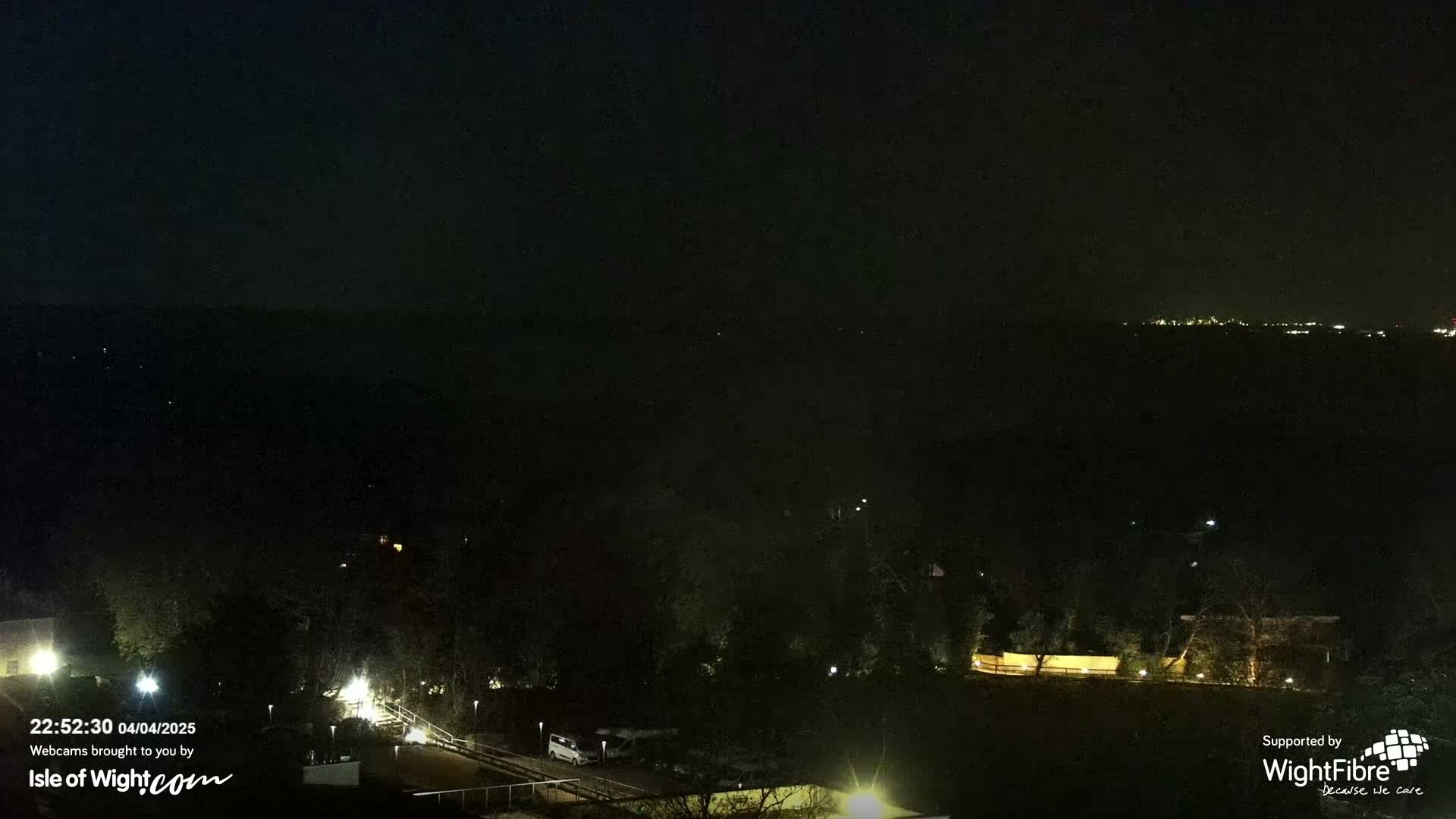 A nighttime, dark view of a landscape with a few scattered lights and distant, faintly visible lights on a horizon.