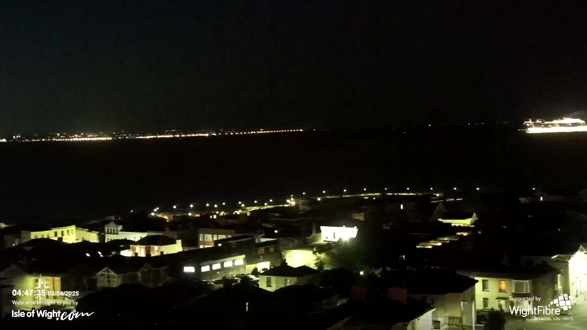A nighttime view of a town overlooking a body of water with a distant line of lights and a large ship on the horizon.