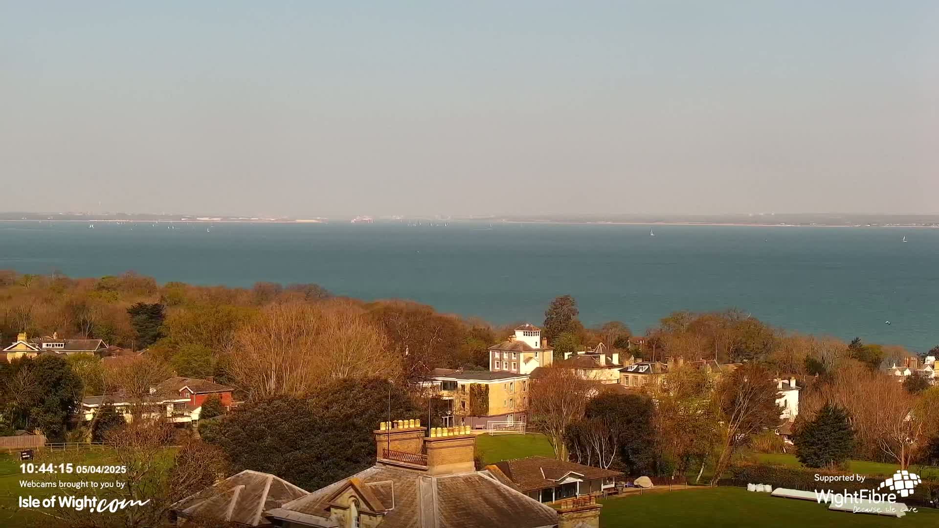 A high-angle, long shot shows a calm body of water with sailboats in the distance, overlooking a tree-lined residential area on a sunny day.