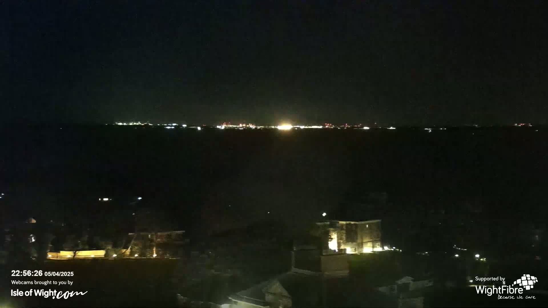 A nighttime long shot shows a distant city skyline glowing faintly on the horizon, with dark, sparsely lit houses and trees in the foreground under a clear, dark sky.