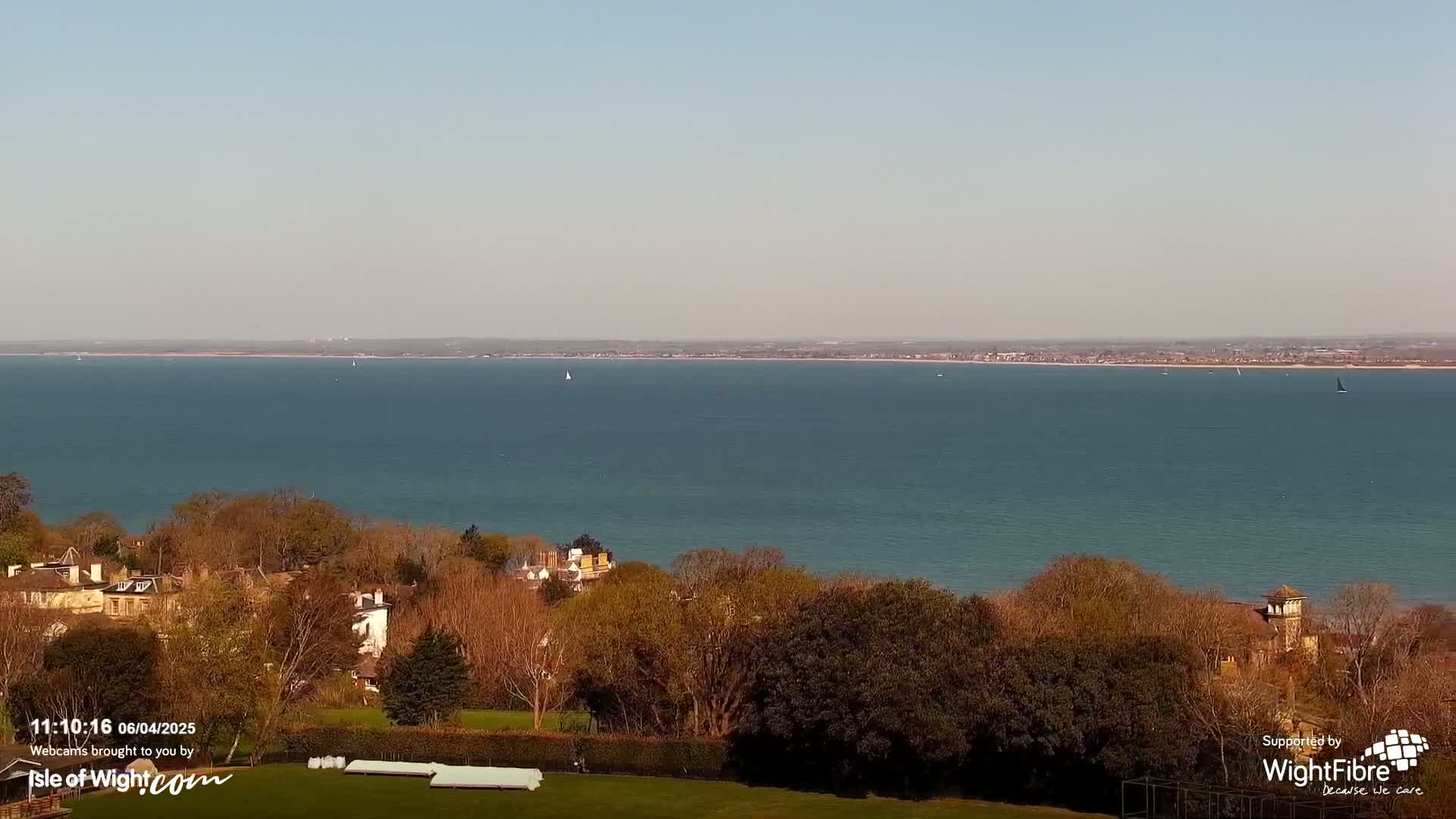 A tranquil coastal scene on a clear day shows a calm expanse of water with several sailboats, overlooking a line of trees and houses on a hillside.