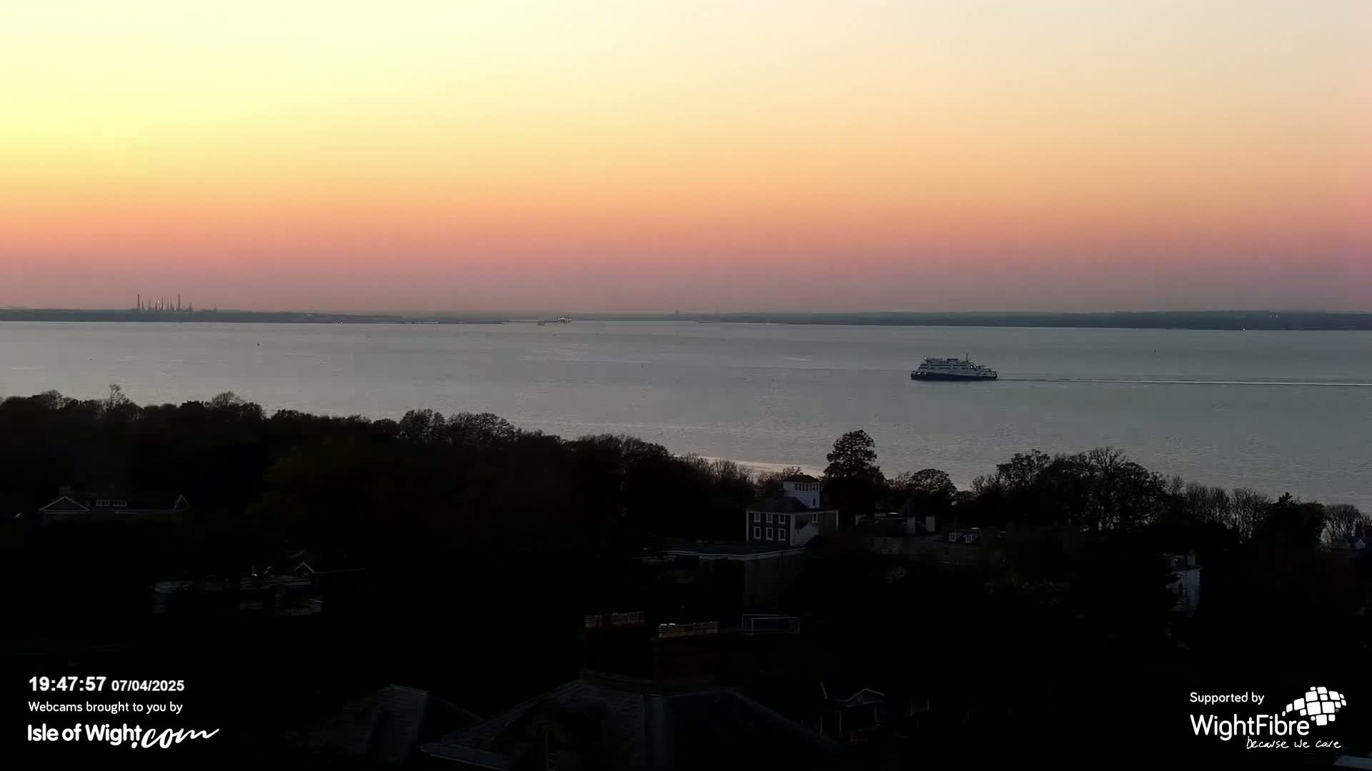 A ferry traverses a calm body of water at sunset, with a shoreline of dark trees and buildings visible in the foreground.