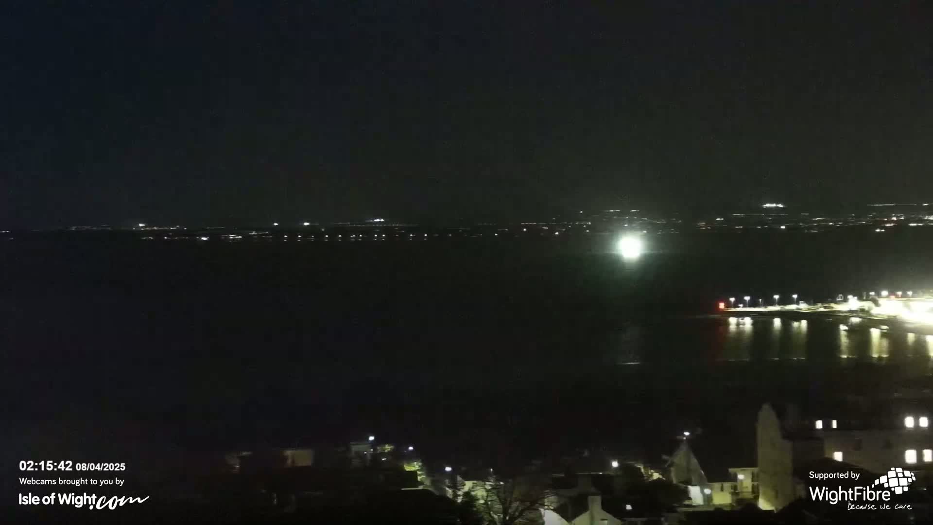 A nighttime view of a dark body of water with faintly lit towns visible across the water and in the foreground.