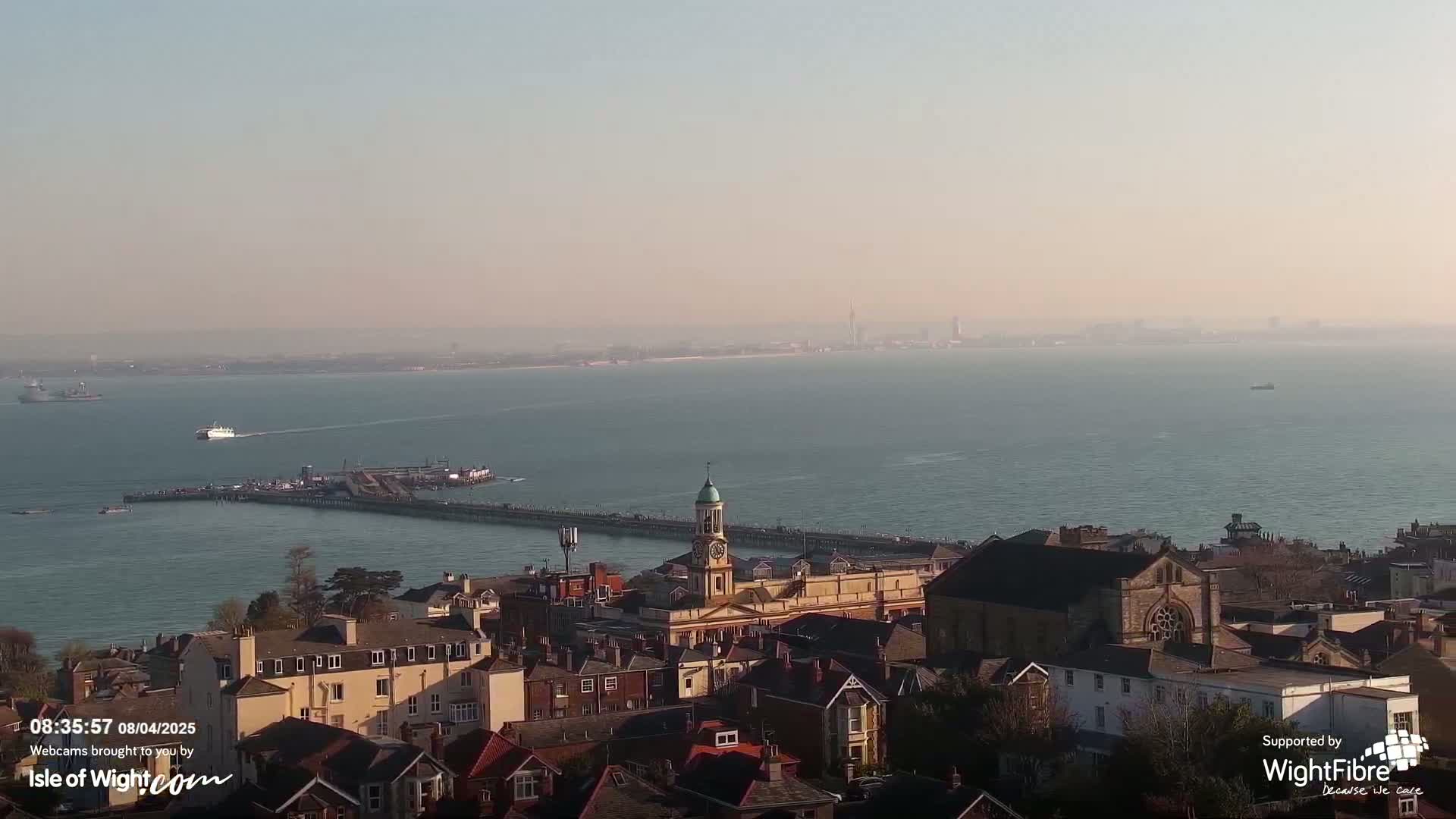 An elevated view of a coastal town on a hazy, sunny day, overlooking a pier and calm ocean waters with several vessels.